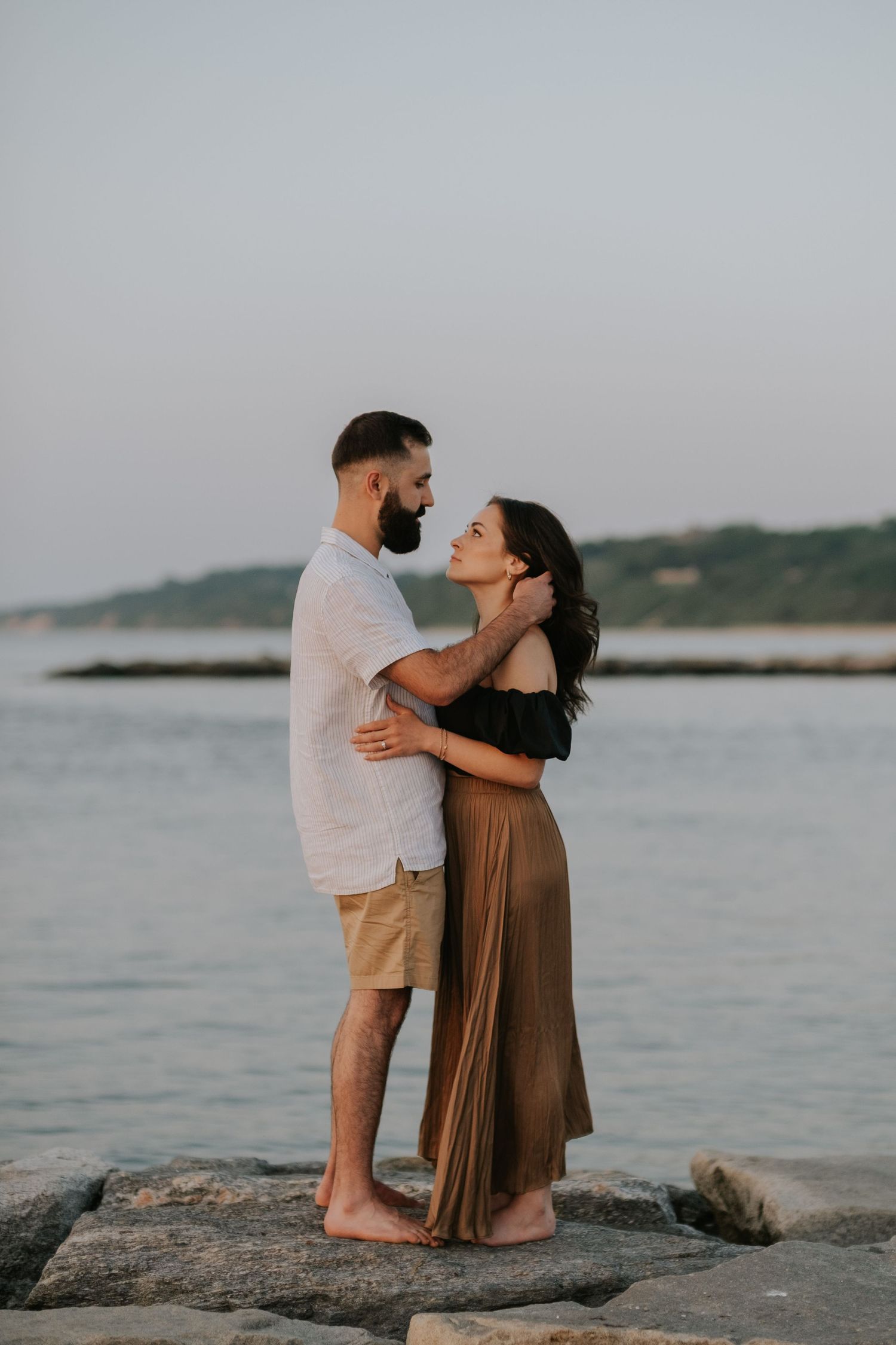 Couple embracing romantically on rocky shore at sunset, man in white shirt and woman in brown dress.