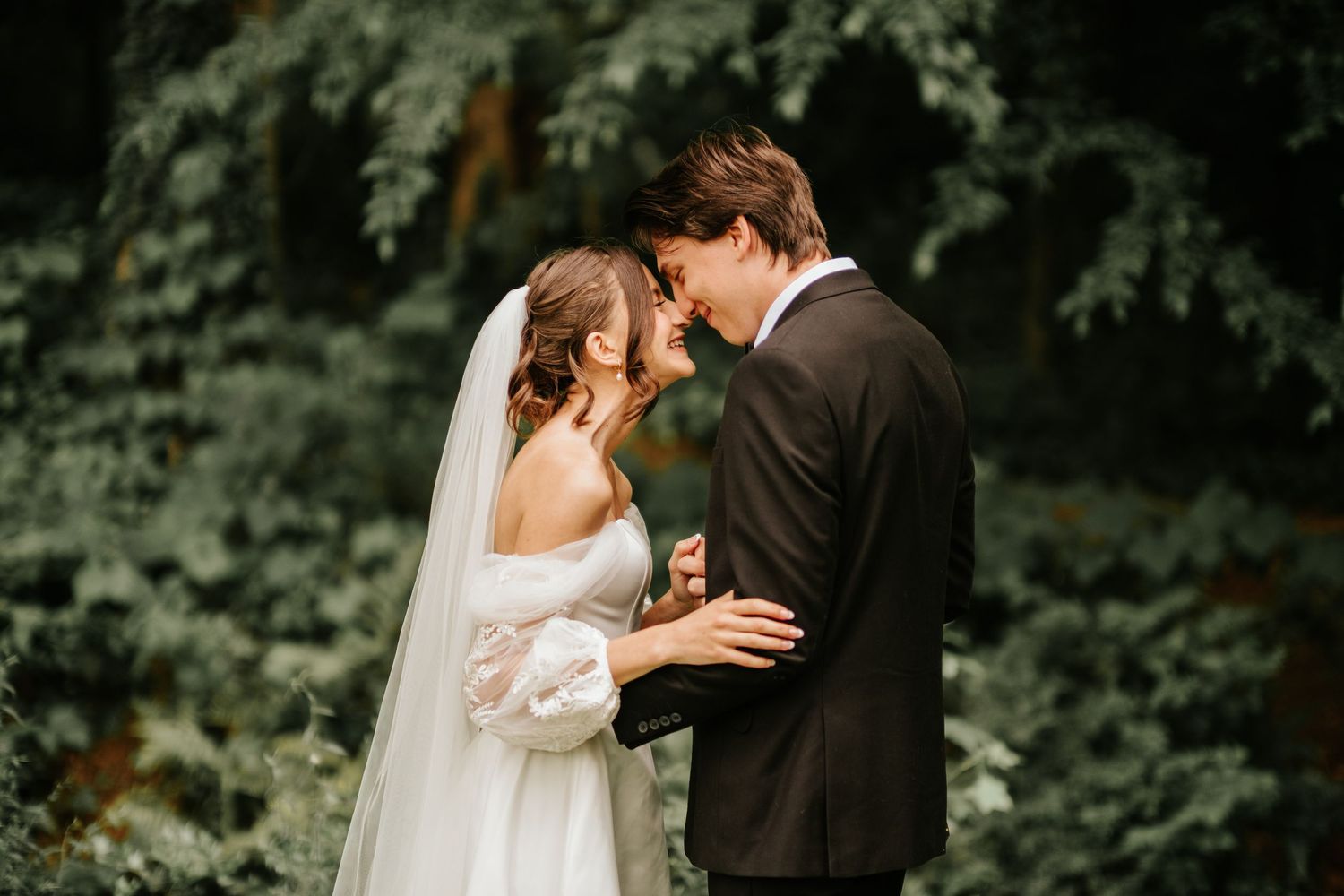 A romantic wedding couple embraces outdoors against a lush green forest backdrop.