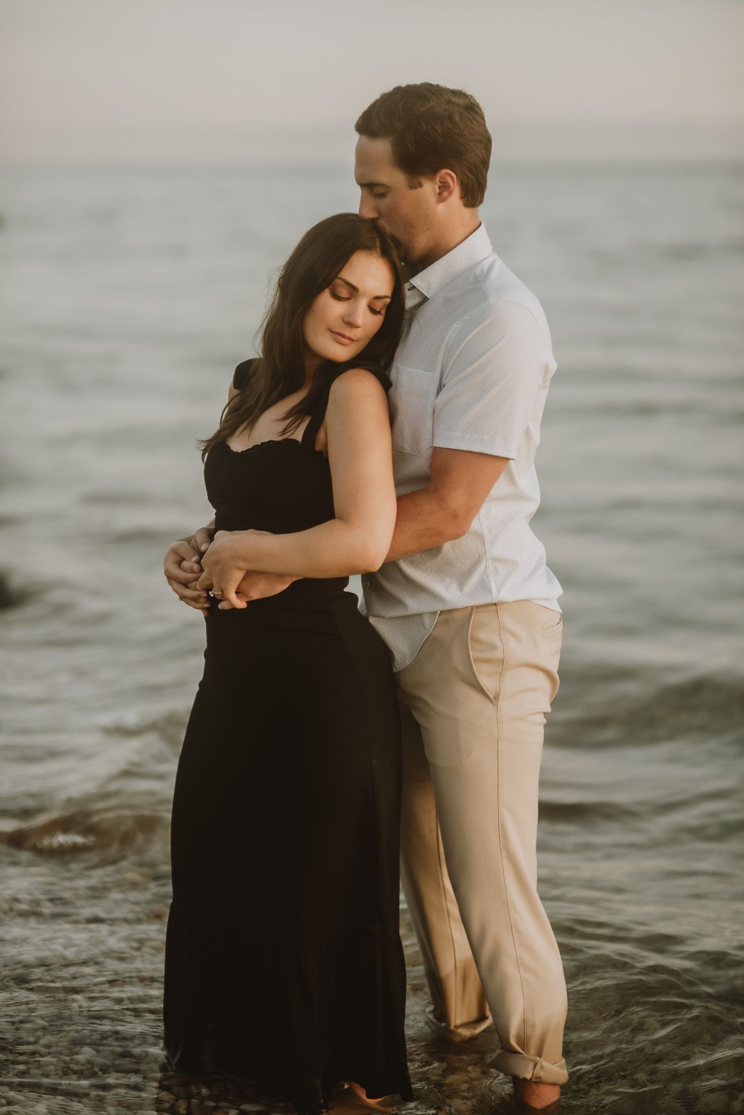 Romantic couple embraces at sunset on beach, with woman in black dress and man in white shirt standing in shallow water.