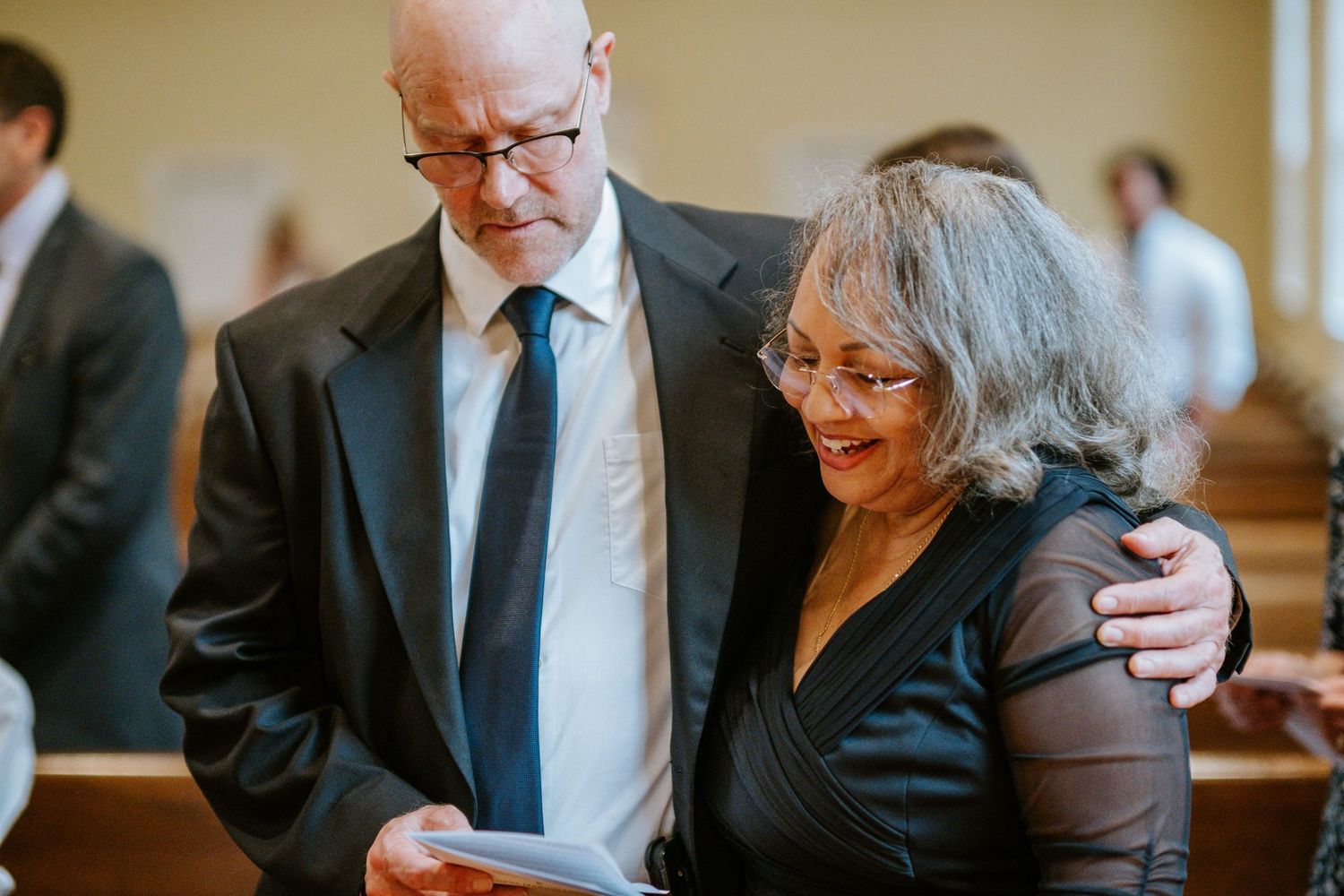 A well-dressed couple in formal attire share an intimate moment while reading together in a church pew.