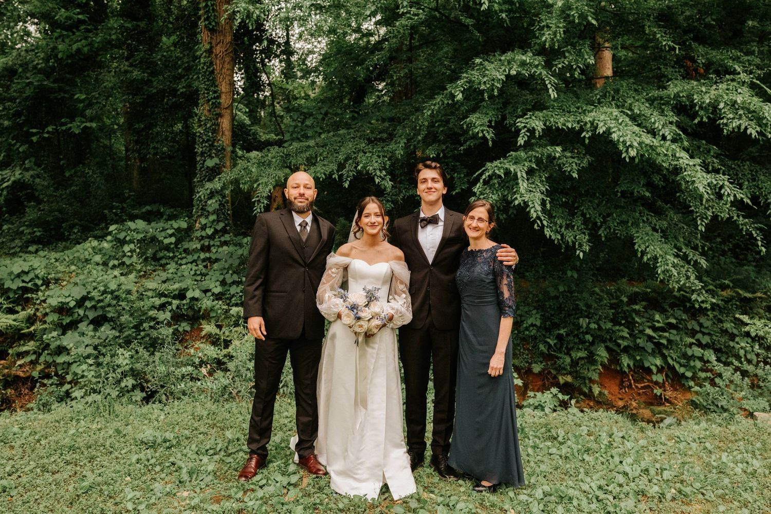 A wedding party of four people poses together outdoors against lush green foliage on a summer day.