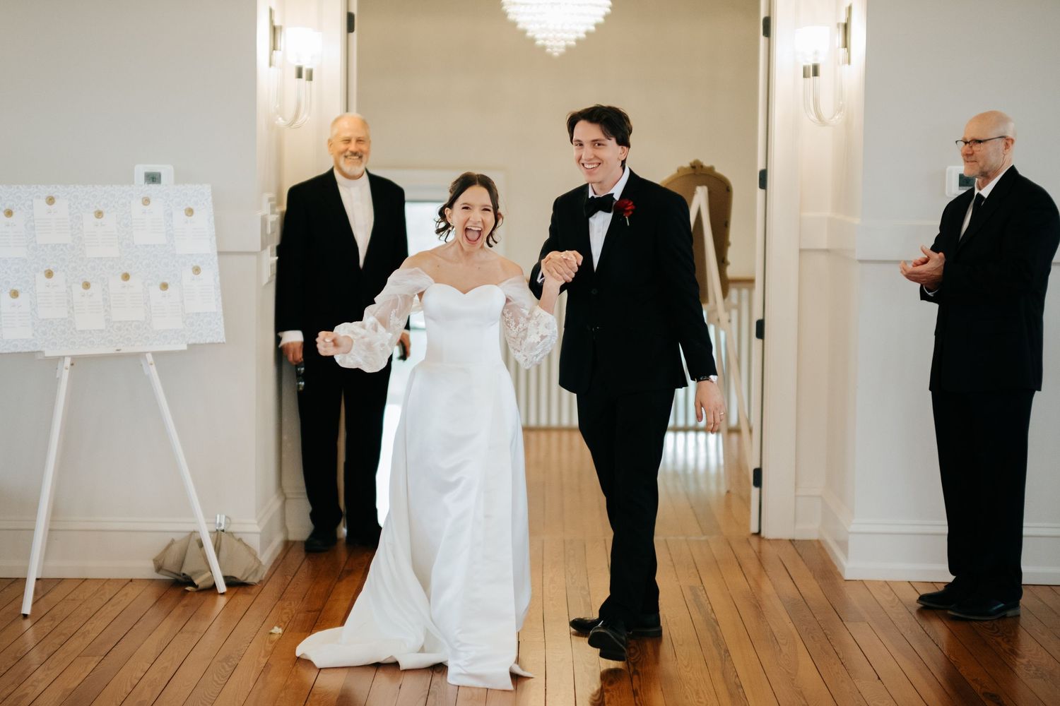 Wedding party members in formal attire gather in an elegant white room with chandelier lighting.