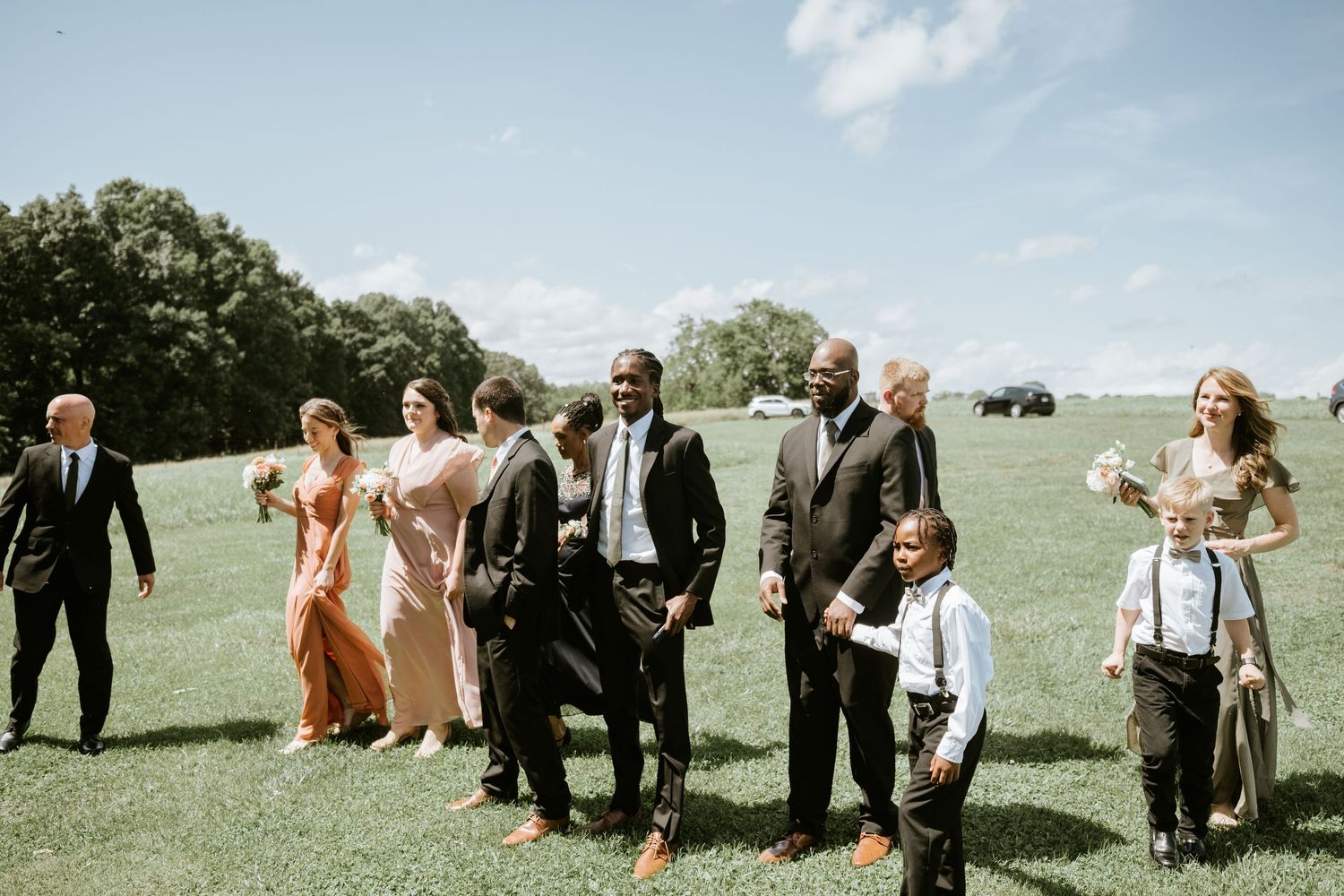 Wedding party walking through a green grassy field on a sunny day with bridesmaids in peach dresses and groomsmen in suits.
