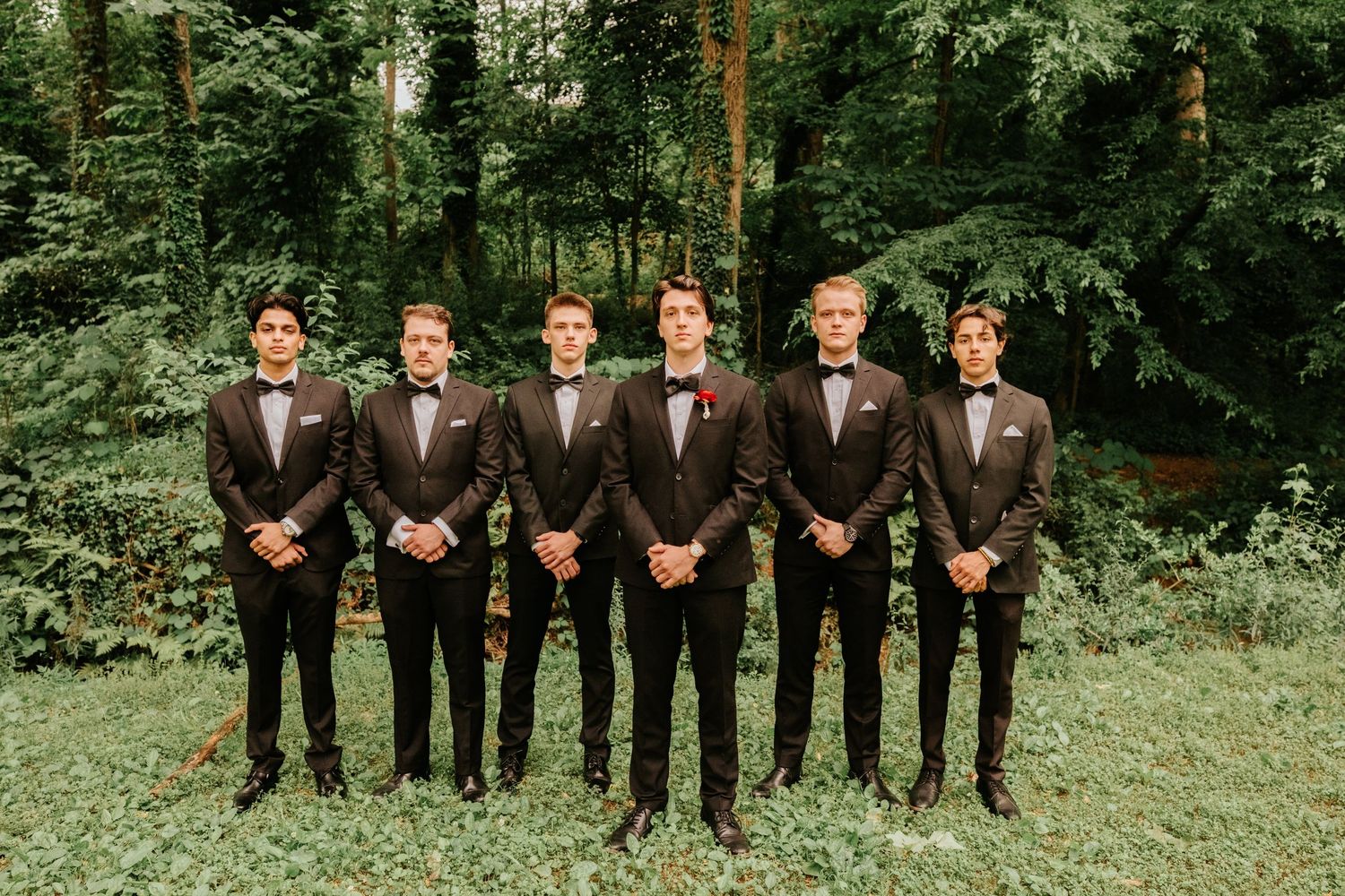 A wedding party in brown suits stands in a line on green grass with trees in the background.