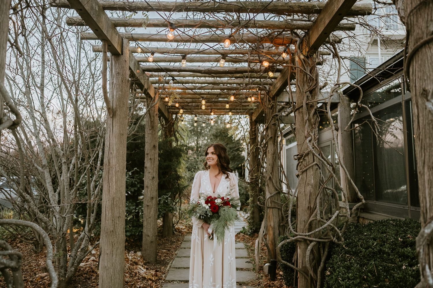 Bride in white gown holding red bouquet stands under rustic wooden pergola with string lights in winter garden.