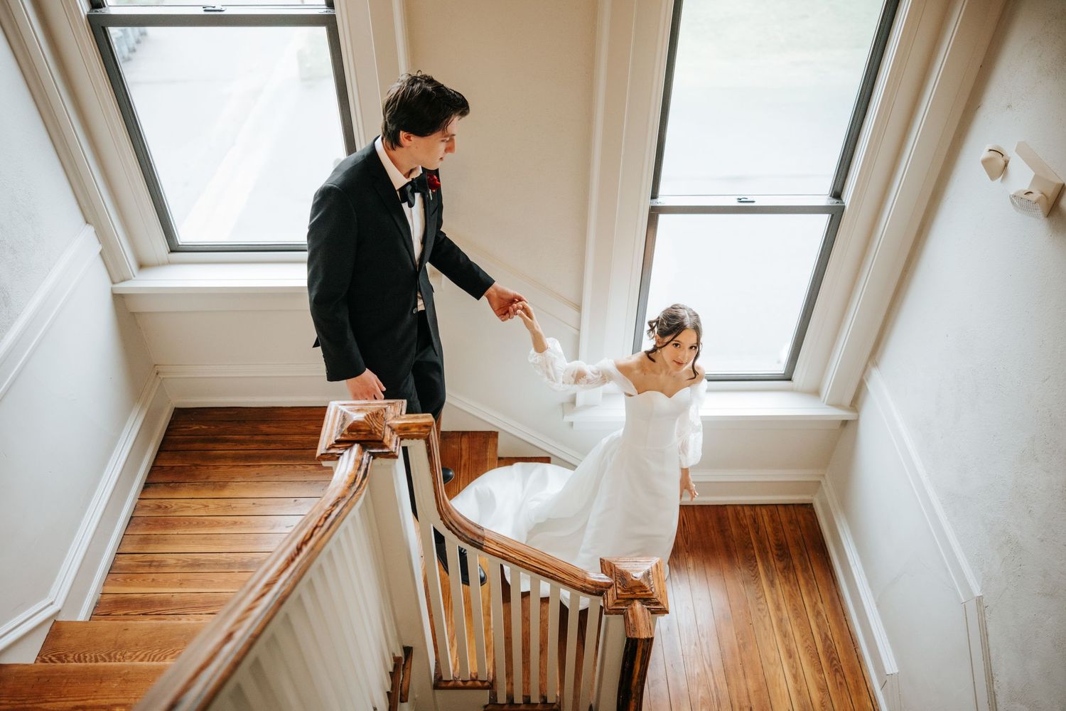A couple descends a wooden staircase in formal wedding attire while holding hands.