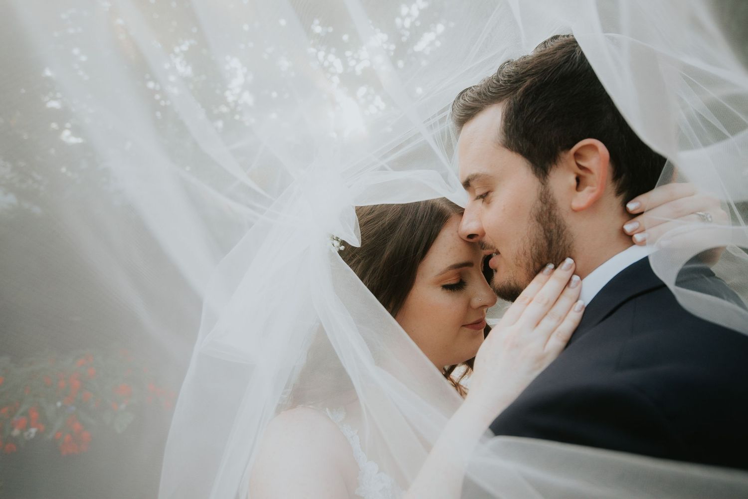 A bride and groom share an intimate moment under a flowing white wedding veil, their foreheads touching tenderly.