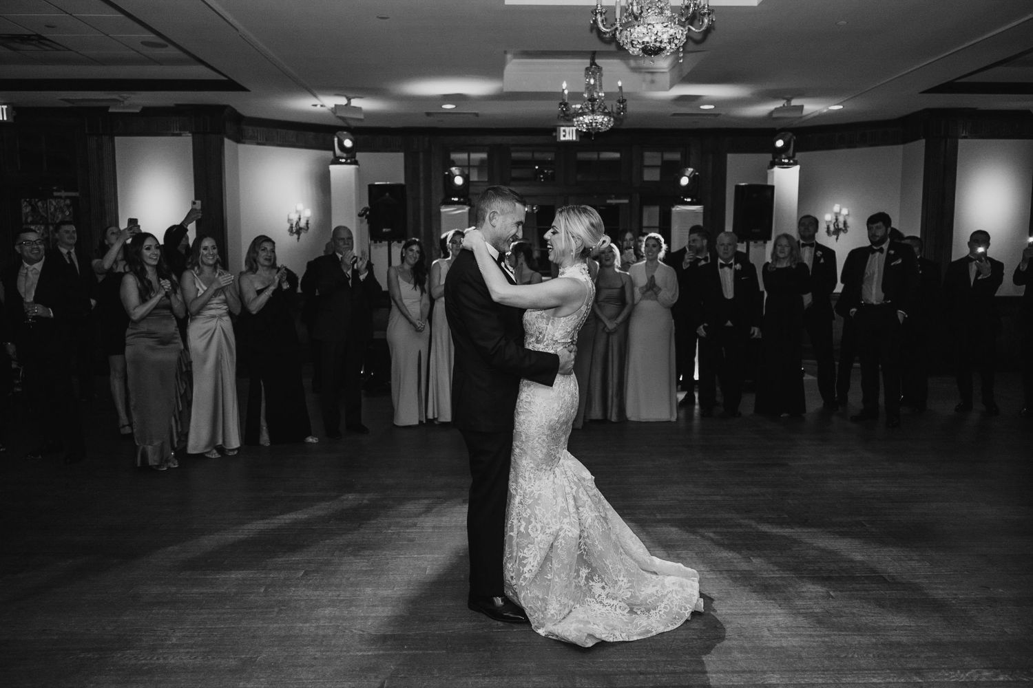 Black and white photo of bride and groom sharing first dance surrounded by wedding guests in elegant ballroom.
