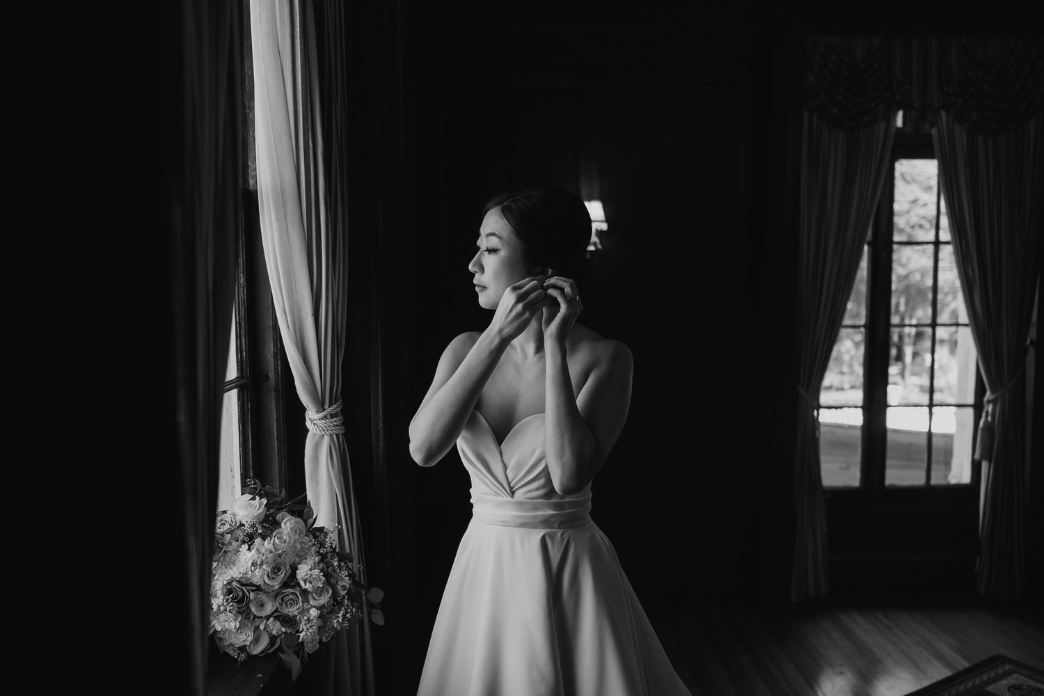 Black and white photo of a bride in a white gown adjusting her earring by a window with soft natural light.