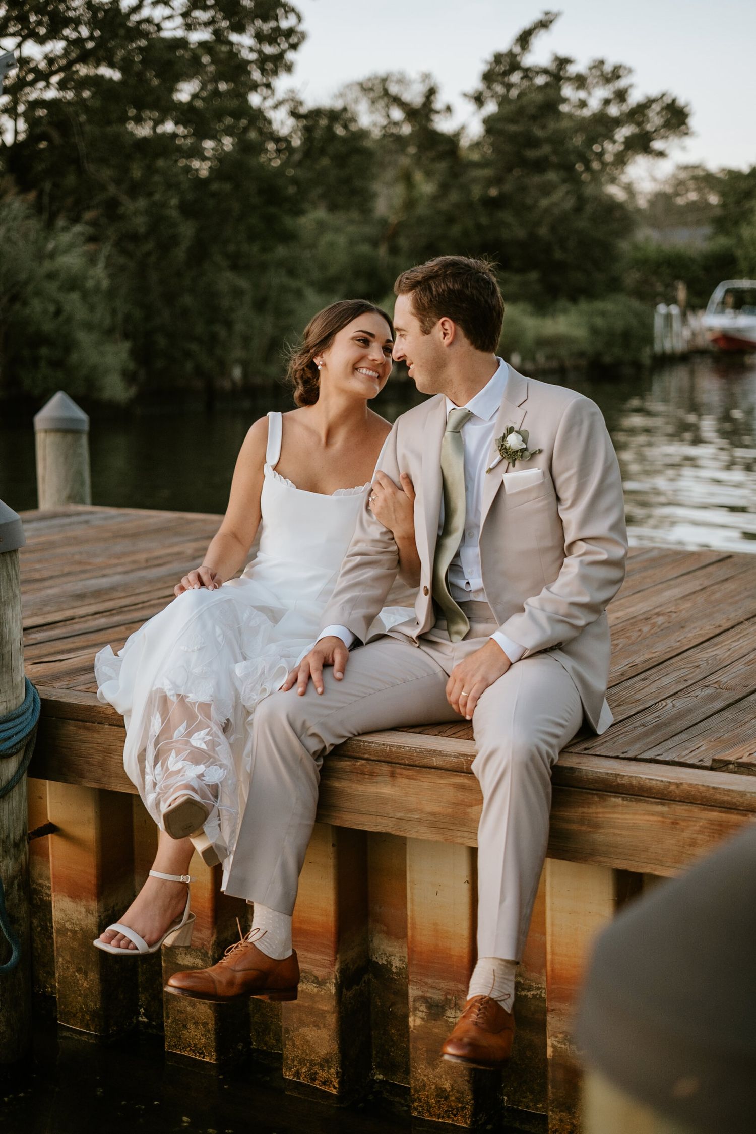 Bride and groom sitting close on a wooden dock by the water, sharing an intimate moment in wedding attire.