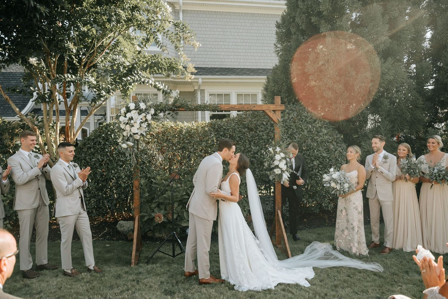 Bride and groom share first kiss at outdoor garden wedding ceremony with bridal party.