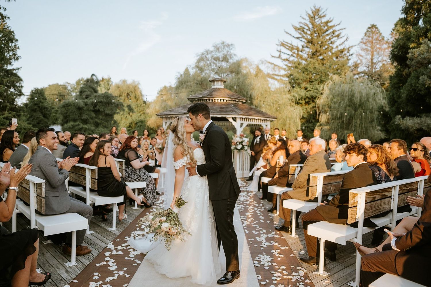 Bride and groom share a kiss at outdoor wedding ceremony with guests cheering, white chairs, and gazebo backdrop.