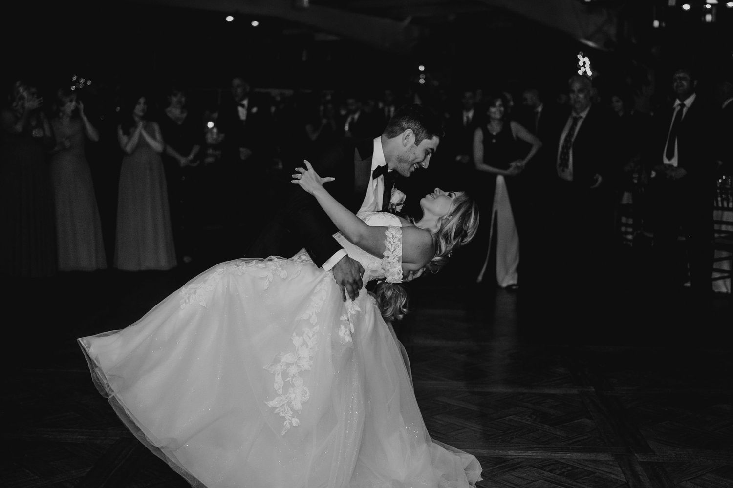 Black and white photo of bride and groom sharing a romantic dip dance surrounded by guests at their wedding reception.