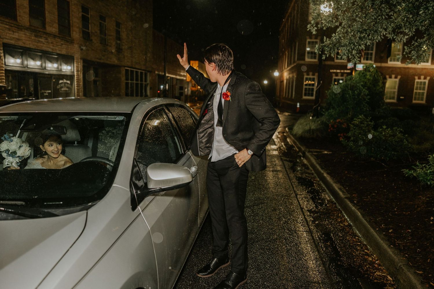 A man in a suit stands by a white car on a rainy night street, illuminated by streetlights.