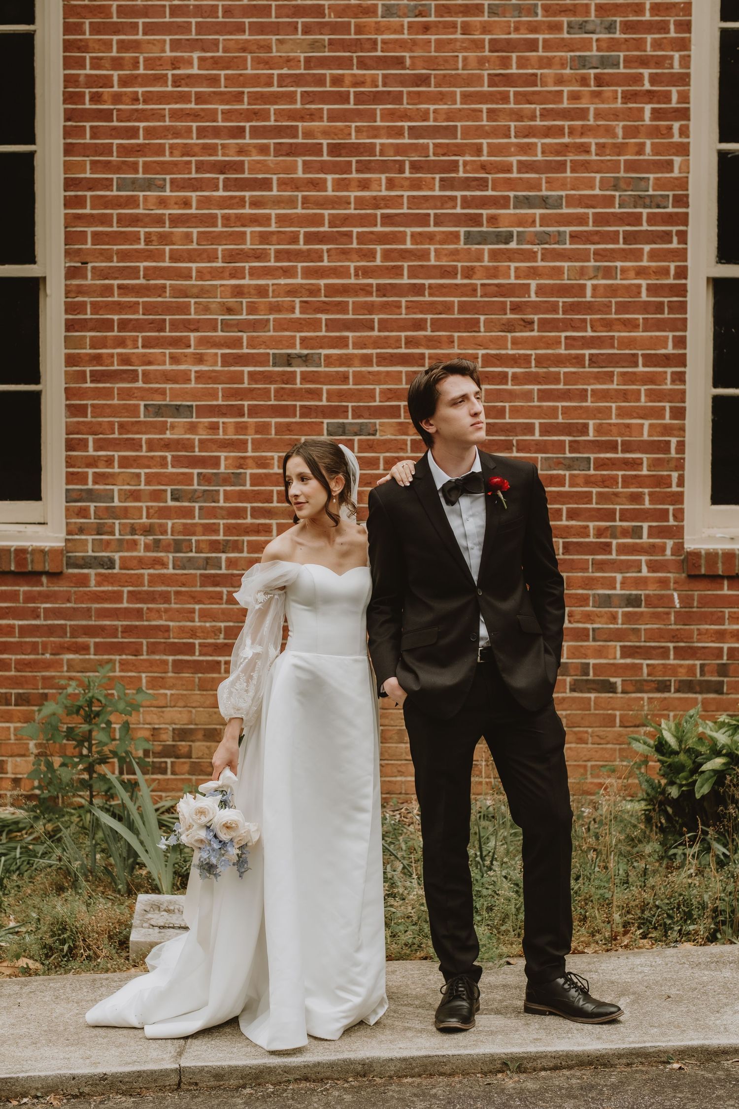 A bride and groom pose casually against a red brick wall during their wedding day.
