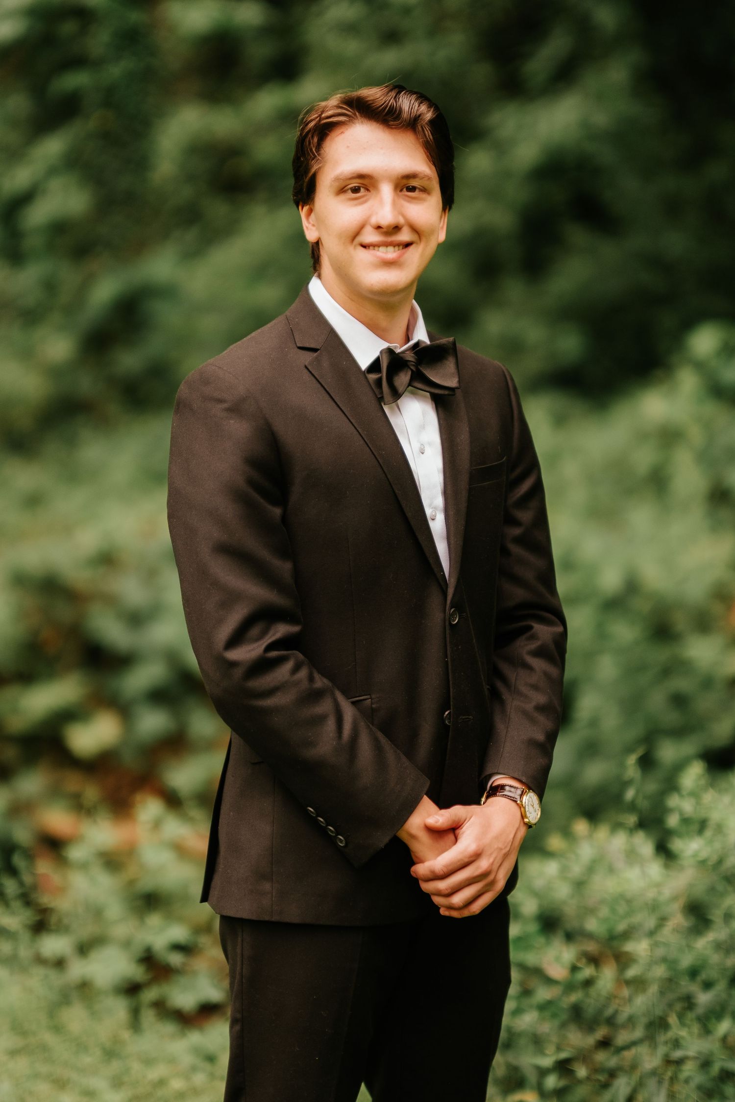 A formal portrait of a person wearing a brown suit and bow tie standing in front of greenery.