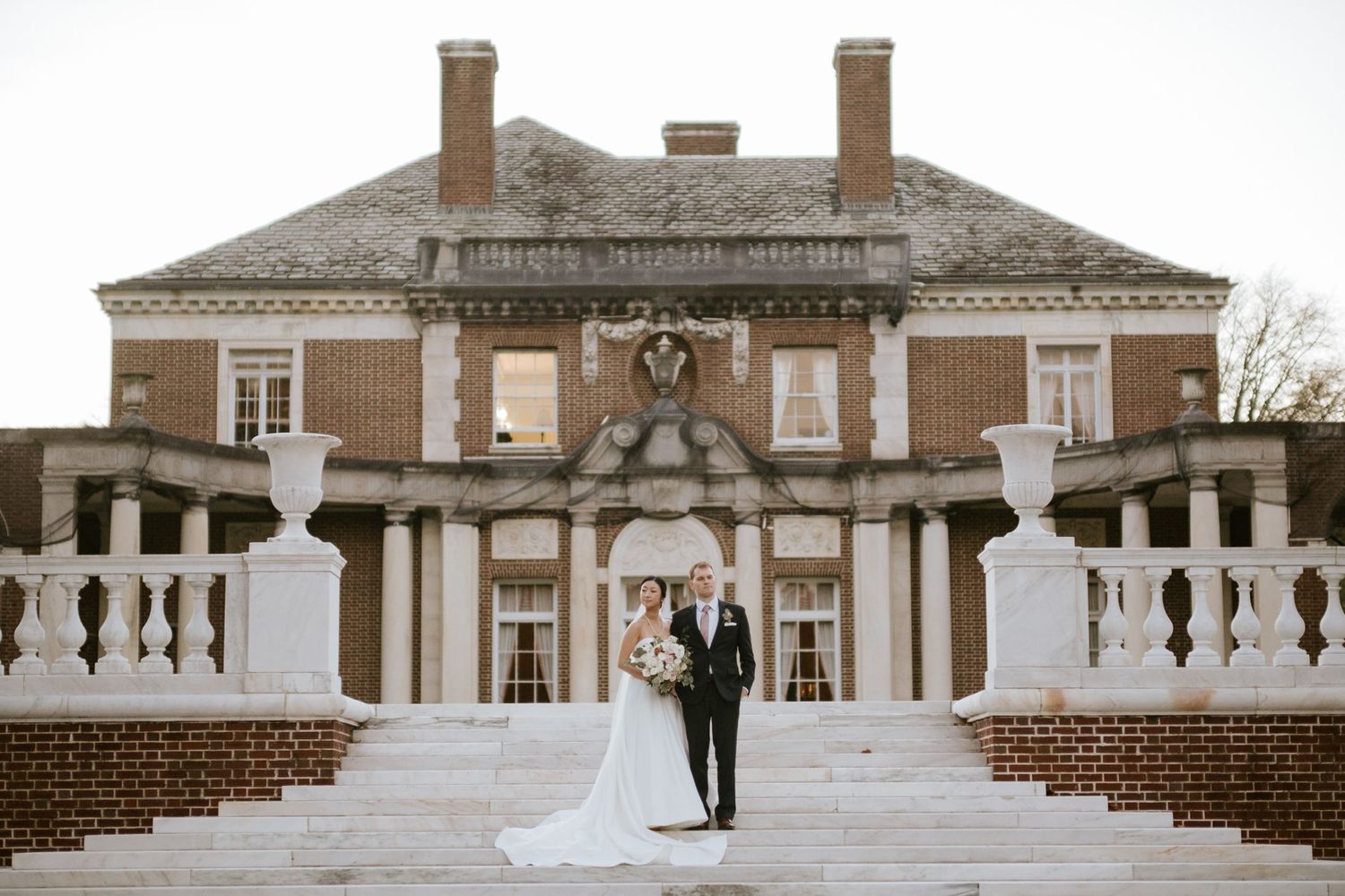 Bride and groom pose on marble steps before an elegant brick mansion at their outdoor wedding ceremony.