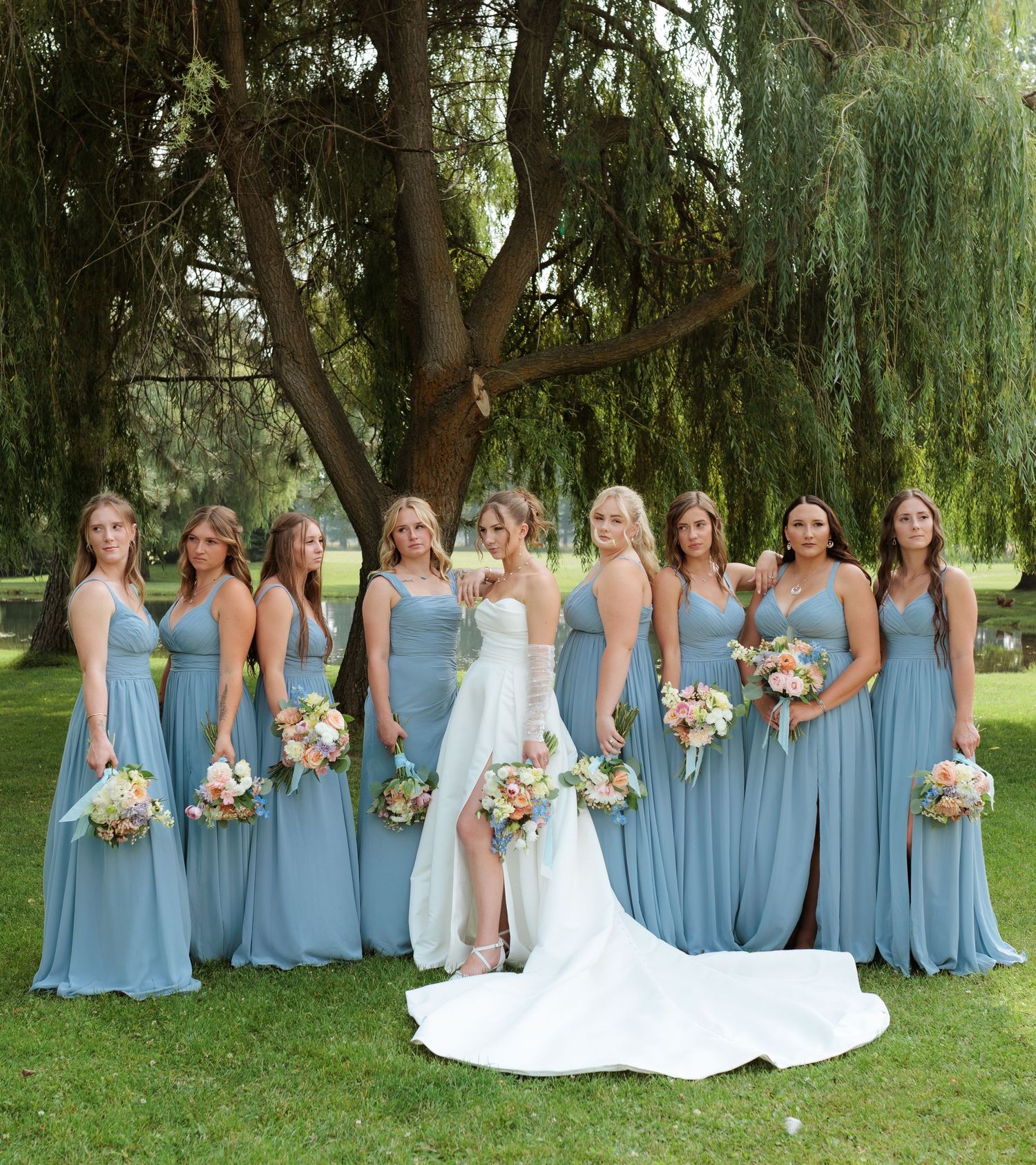 Bride in white gown poses with eight bridesmaids in dusty blue dresses holding colorful bouquets under a willow tree in Montana.