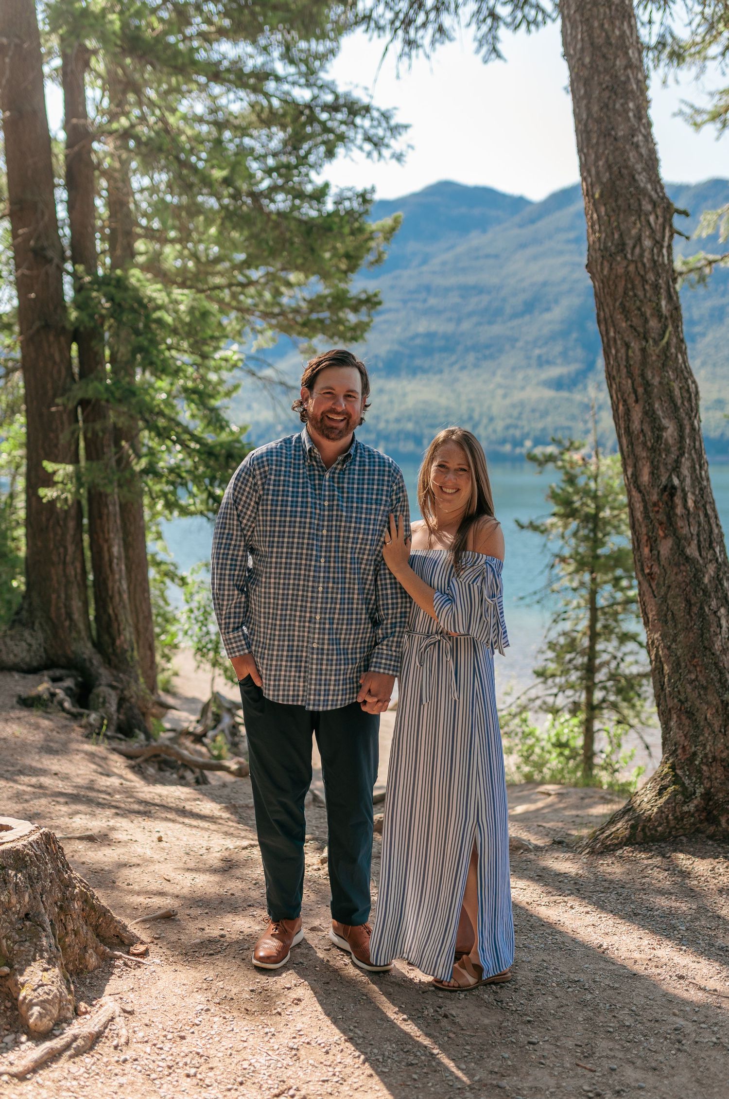 A couple poses together on a mountain trail overlooking a scenic lake surrounded by evergreen trees.