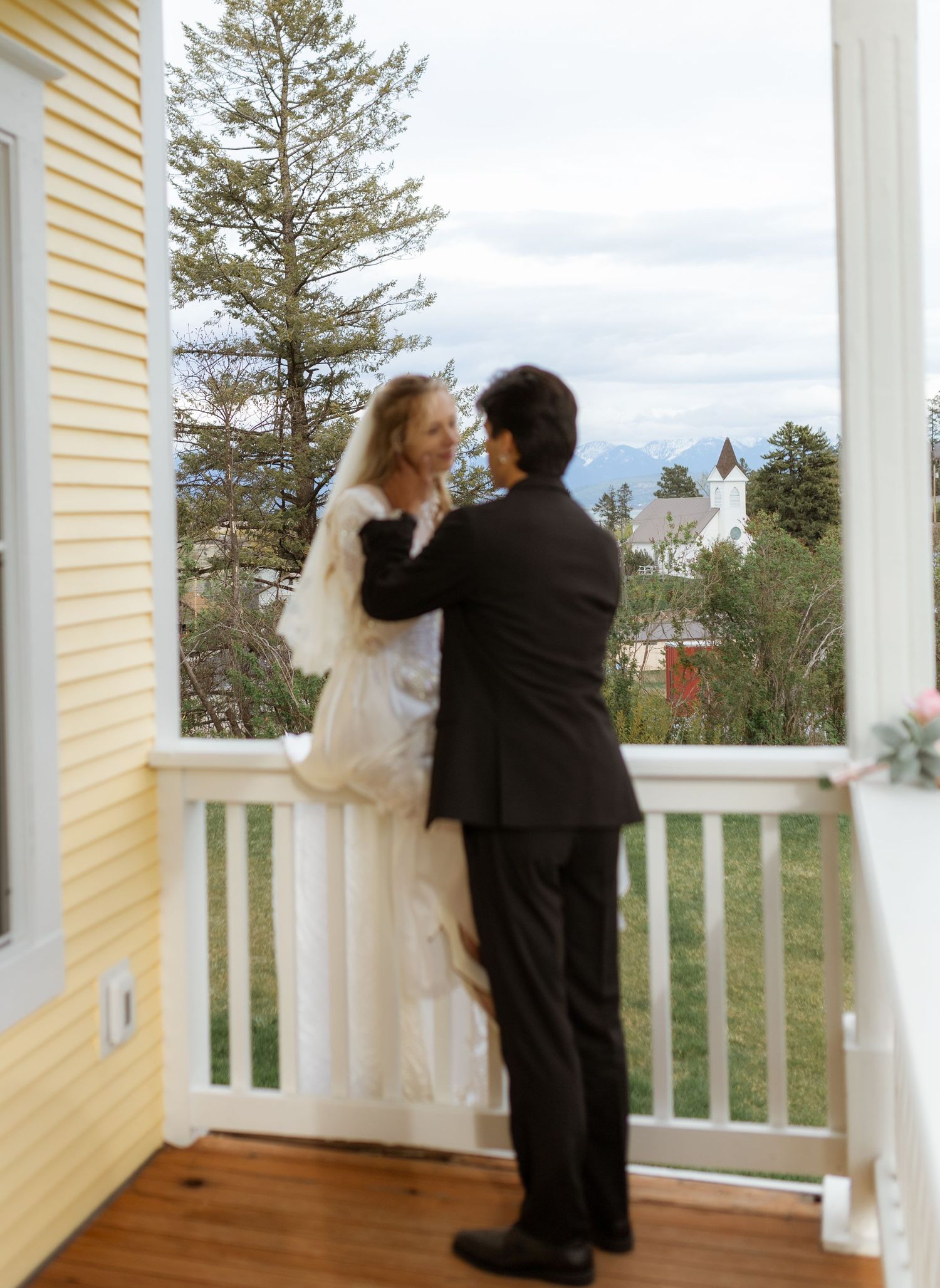 A couple stands on a white porch overlooking a scenic view with mountains and a church steeple visible in the distance.