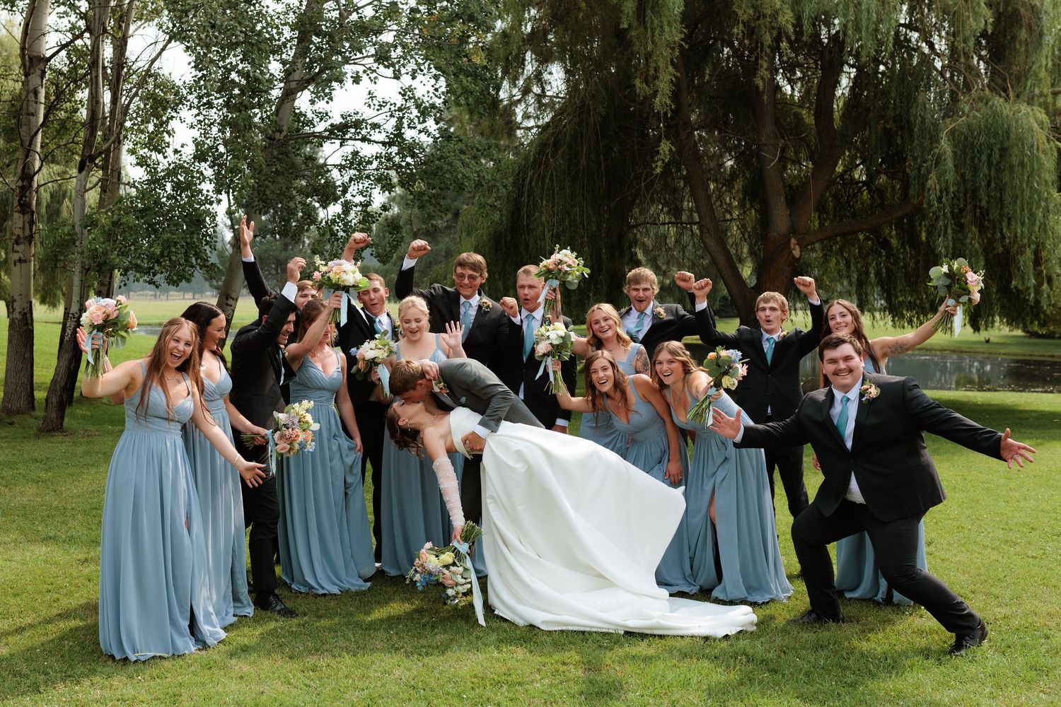 Wedding party celebrating outdoors, bridesmaids in blue gowns and groomsmen cheering under willow trees while the bride and groom kiss.