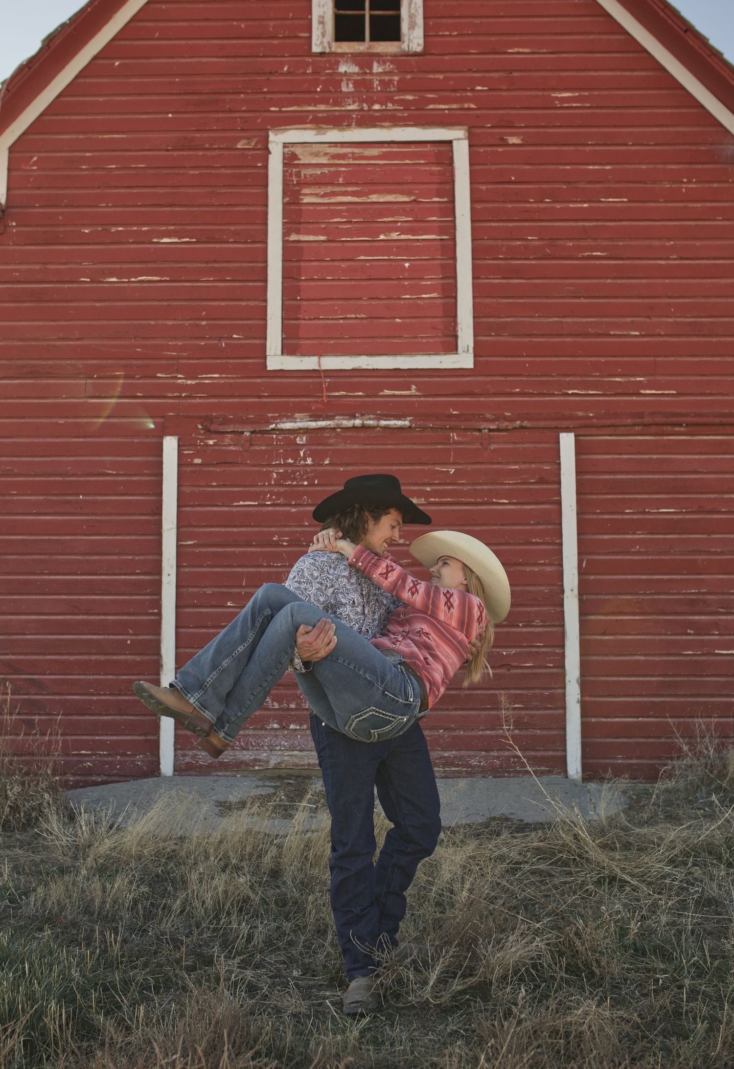 A romantic couple embraces in front of a rustic red barn with weathered siding and a white framed window.