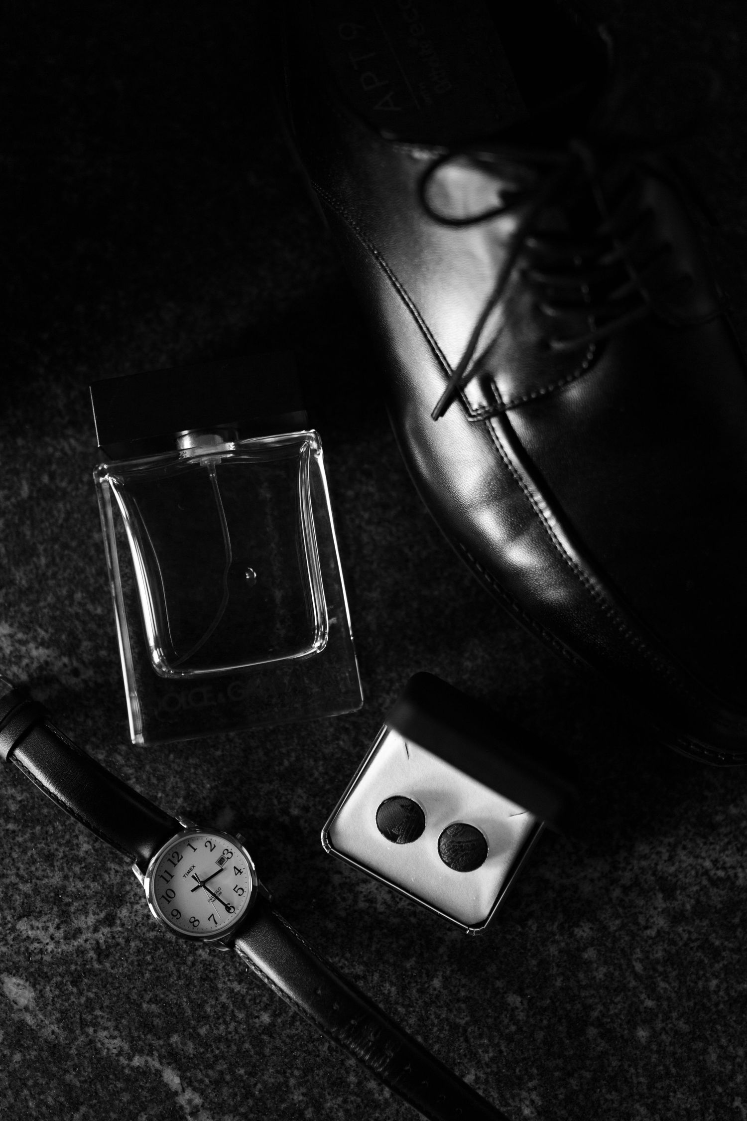 Black and white flat lay of groom's wedding details including dress shoes, cologne, watch, cufflinks, and belt on dark surface in Montana.