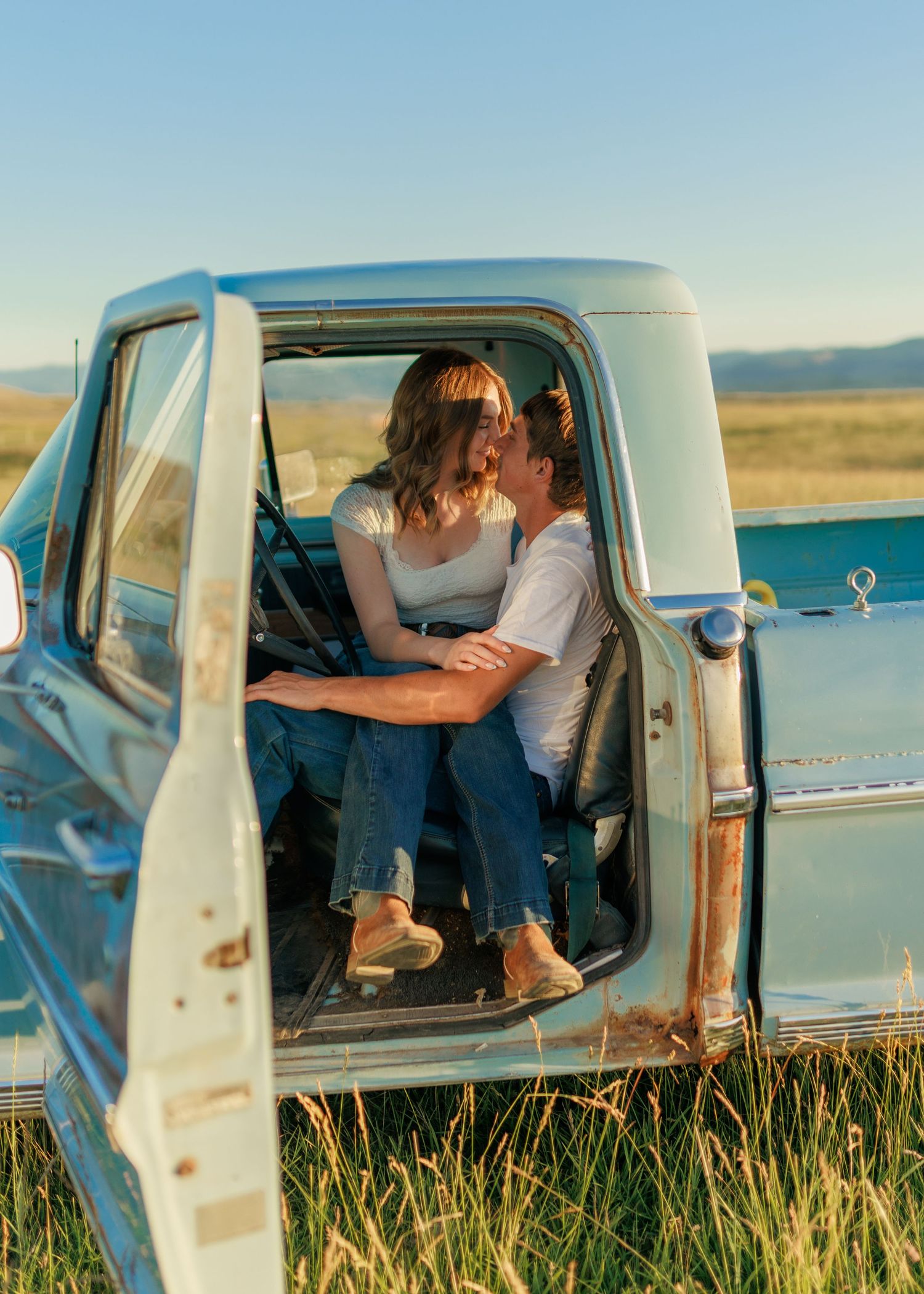 Couple sharing a romantic moment inside a vintage blue pickup truck in a golden field at sunset.