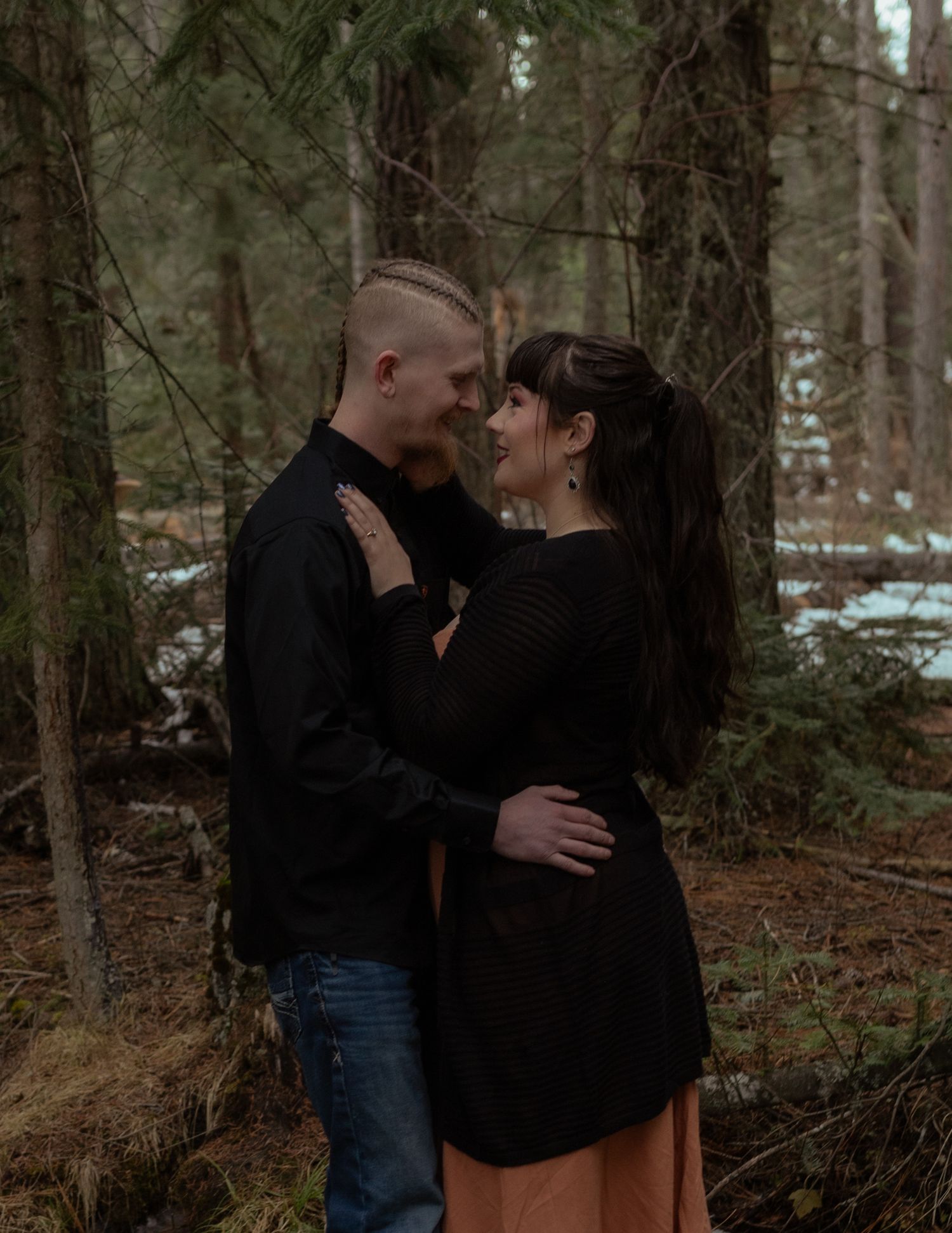 A couple embraces in an intimate moment surrounded by tall evergreen trees in a forest.