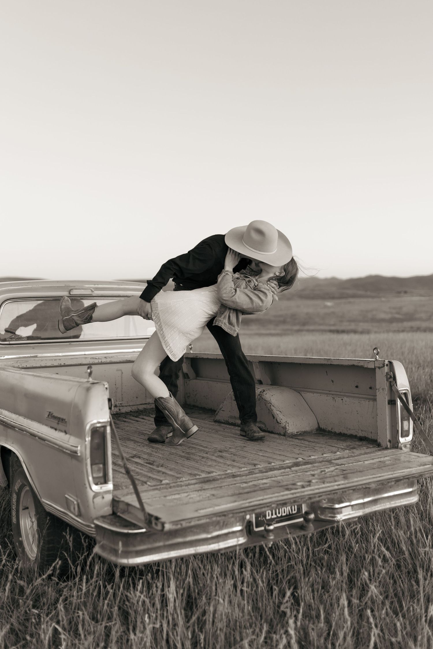 Two people relax on the bed of a vintage pickup truck in a black and white rural landscape.