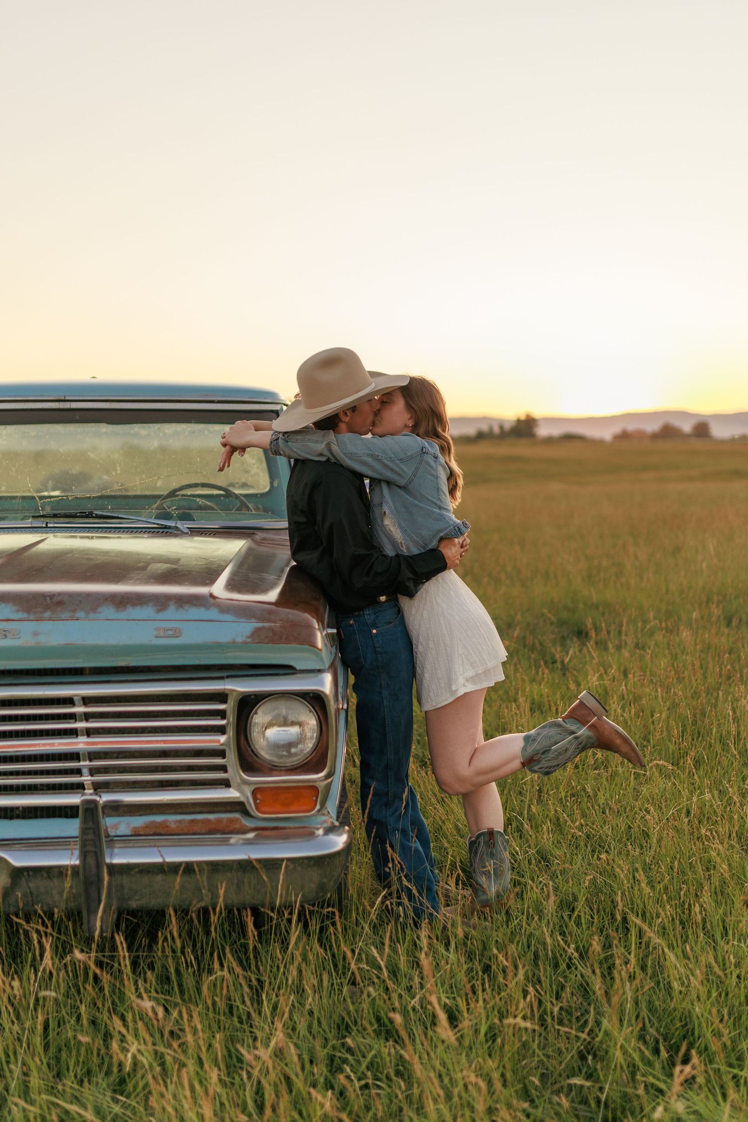 Couple in western attire sharing romantic moments by classic car in grass field.