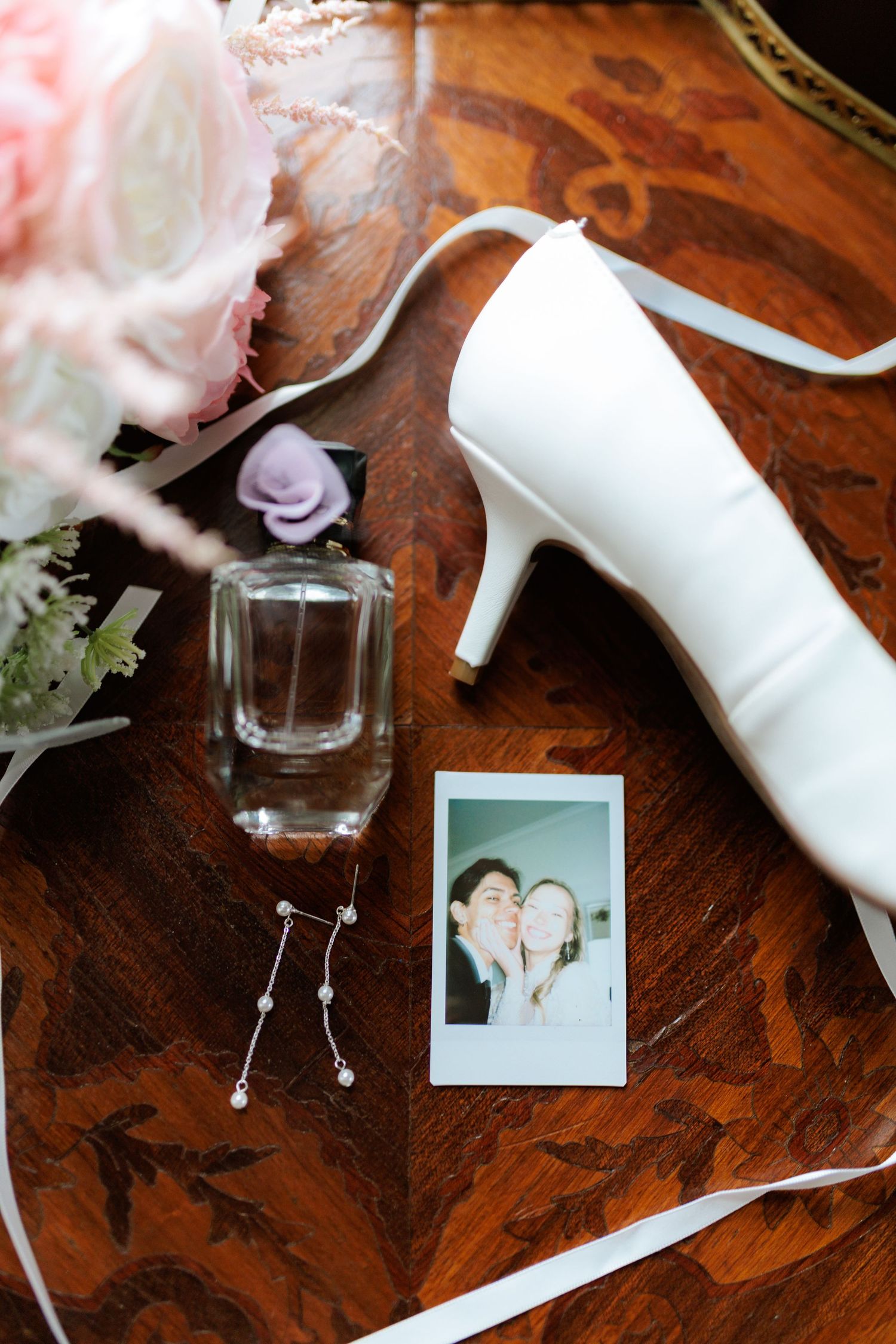 Bridal flat lay with white heels, perfume, pearl earrings, Polaroid photo, and pink floral bouquet on wood table in Montana.