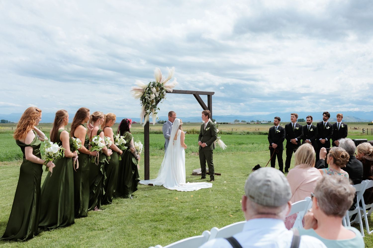 Montana outdoor wedding ceremony on green field with bridesmaids in olive dresses, groomsmen in black, under rustic wooden arch and mountain