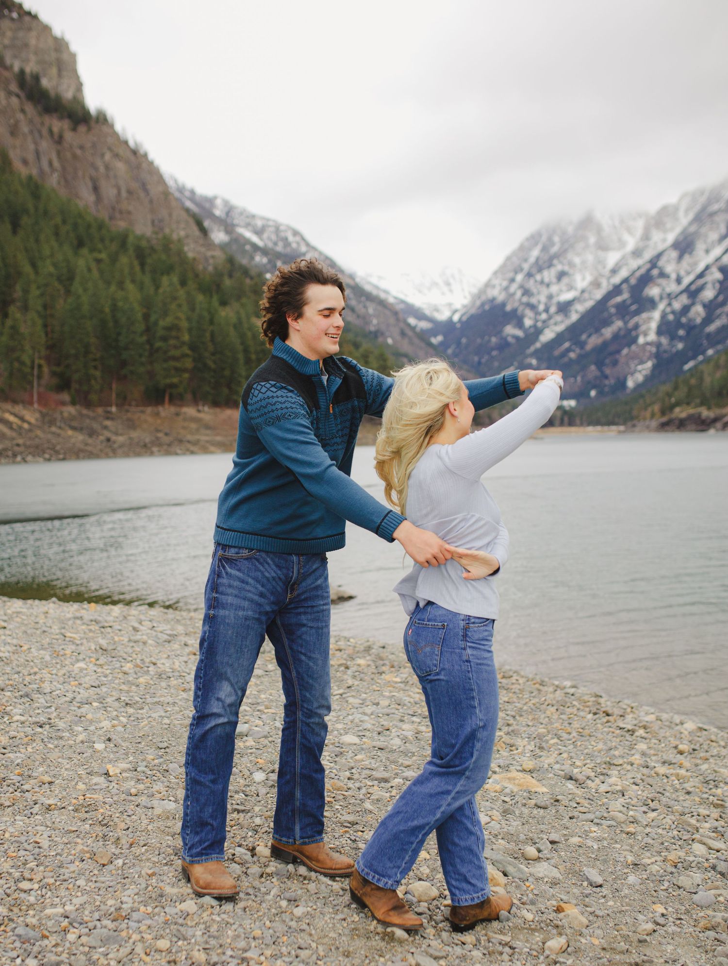 Playful couple dancing on rocky shore with mountain lake backdrop.