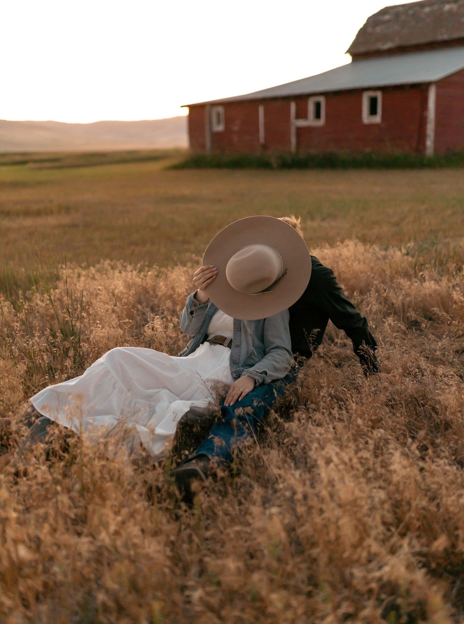 Person in a wide-brimmed hat sitting in a golden field near a rustic red barn at sunset.