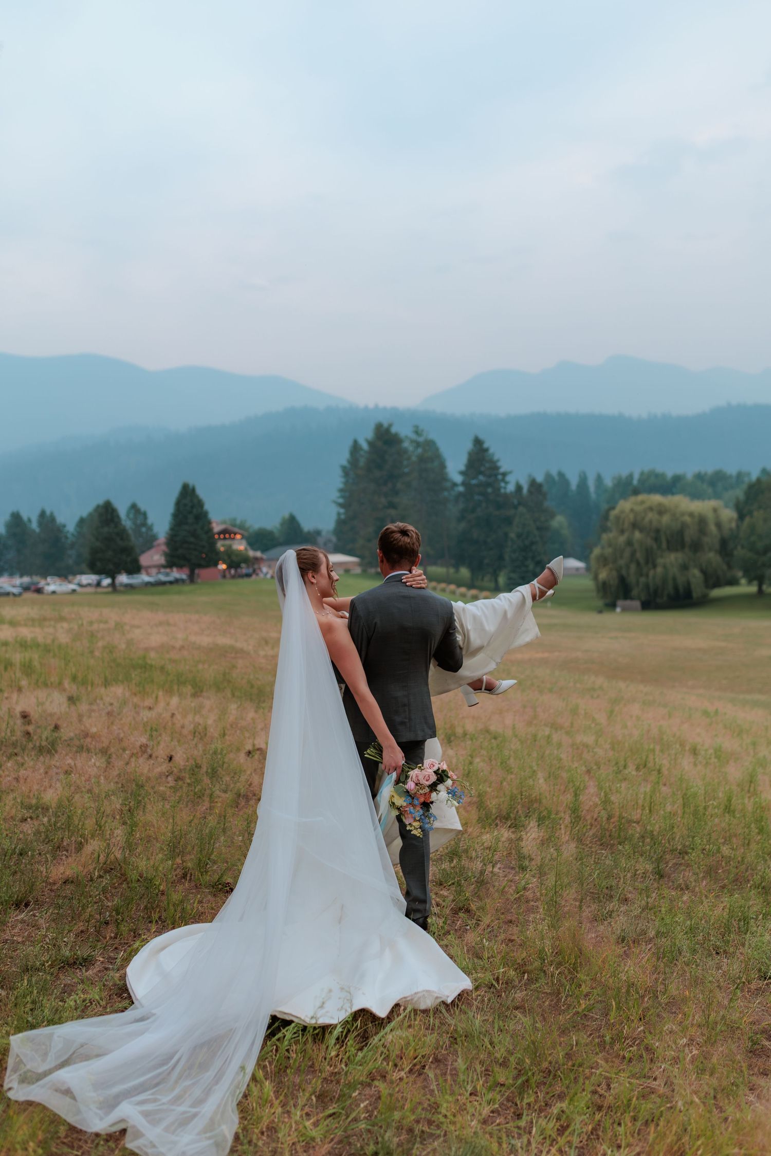 Bride in a long flowing veil and wedding gown embraces her groom in a gray suit in a grassy meadow with misty mountains behind.