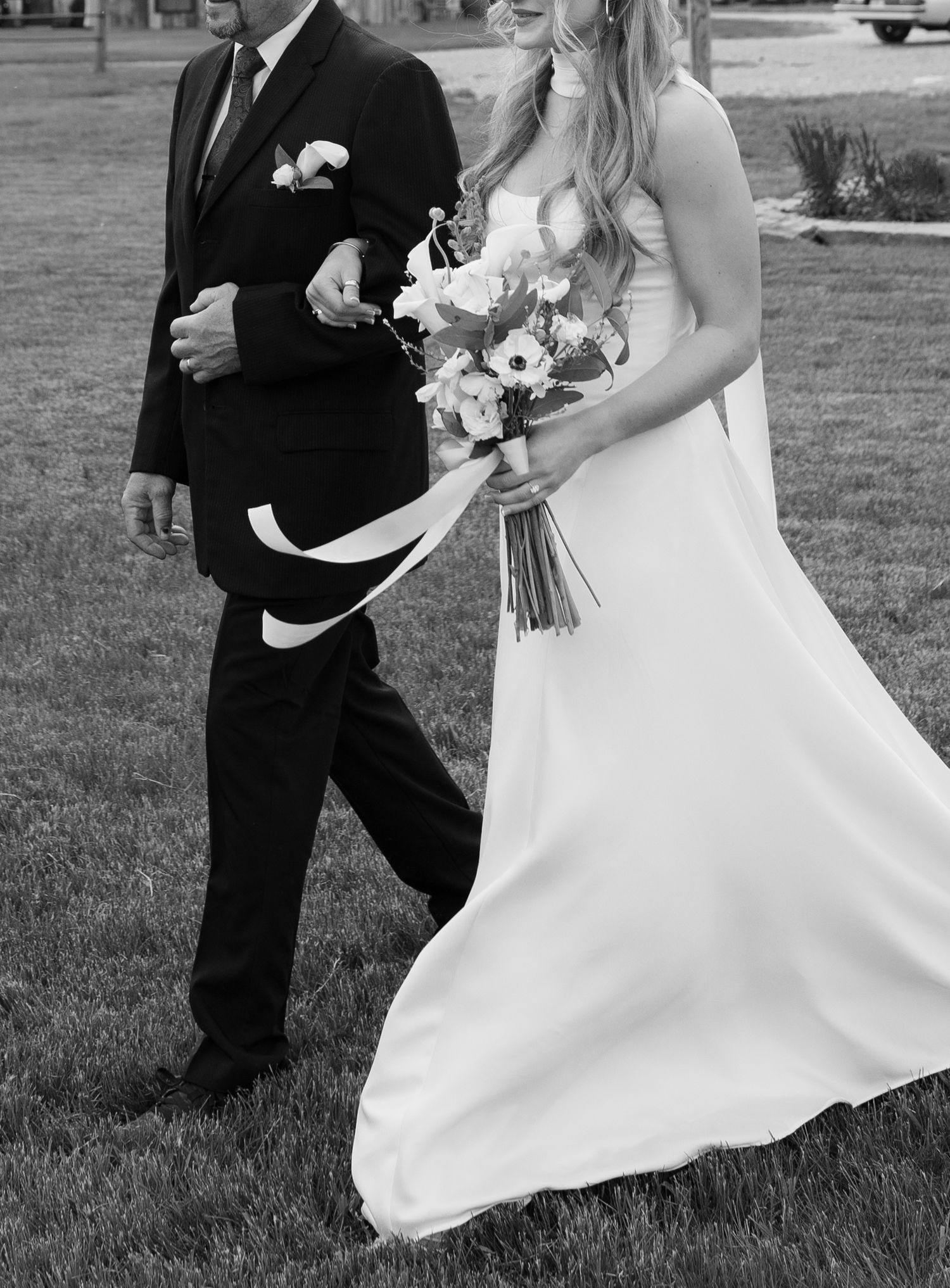 Black and white photo of a bride holding a bouquet walking arm in arm with her dad at Montana ceremony with mission mountain views