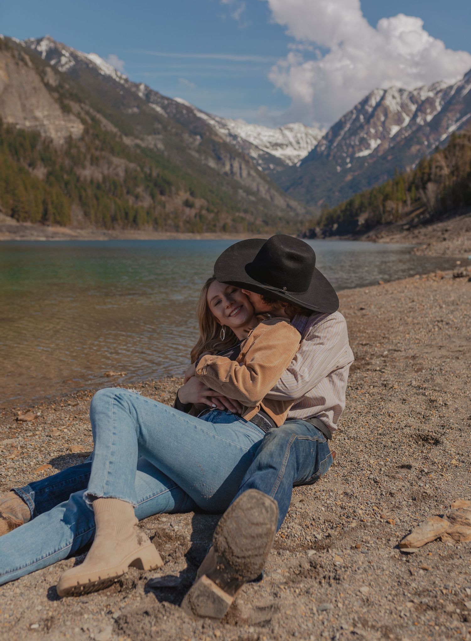 A couple in casual wear relaxes on a rocky beach with mountains in the background.