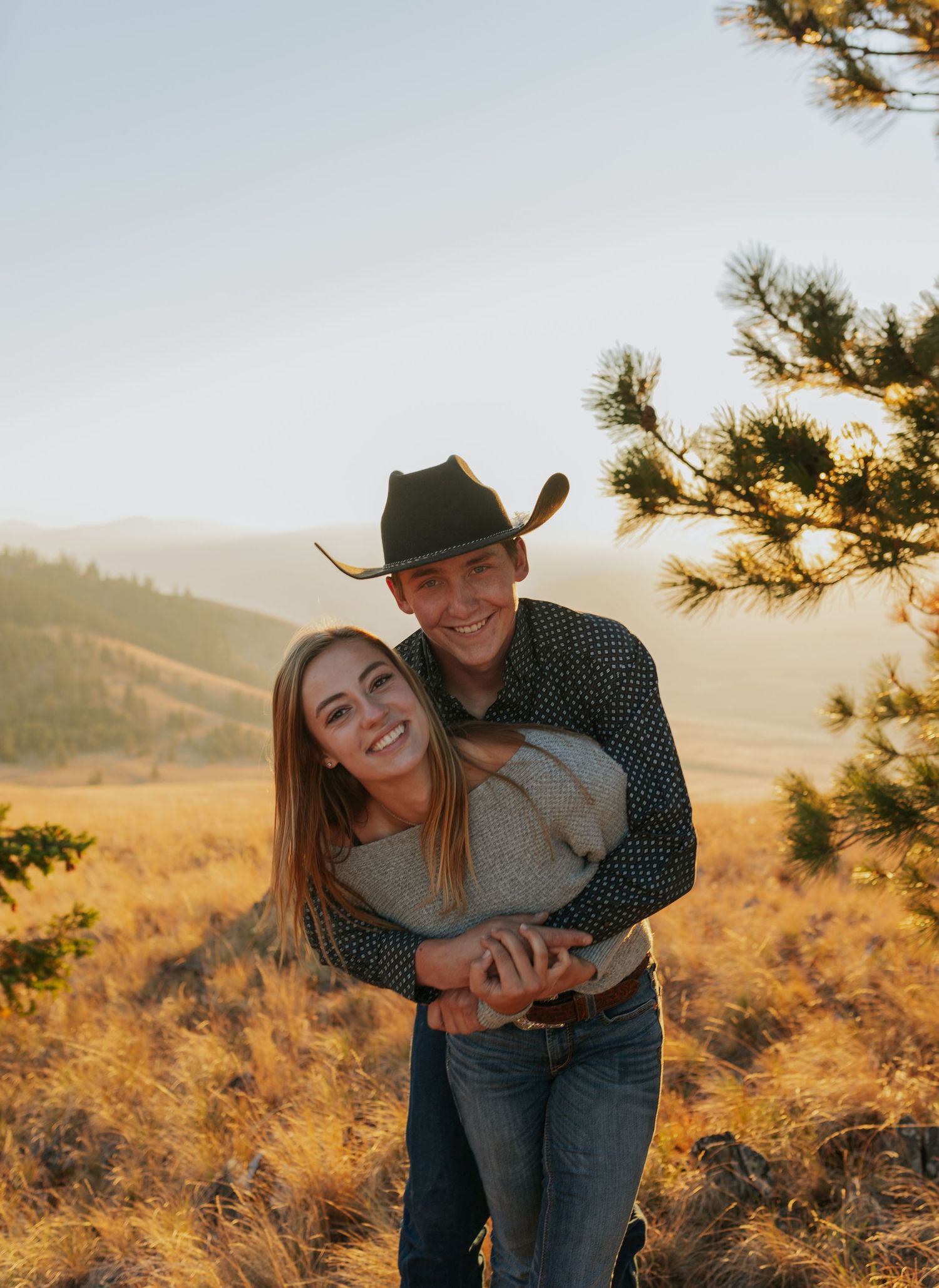 Romantic couple embraces in a golden sunset field with mountains and pine trees in the background.