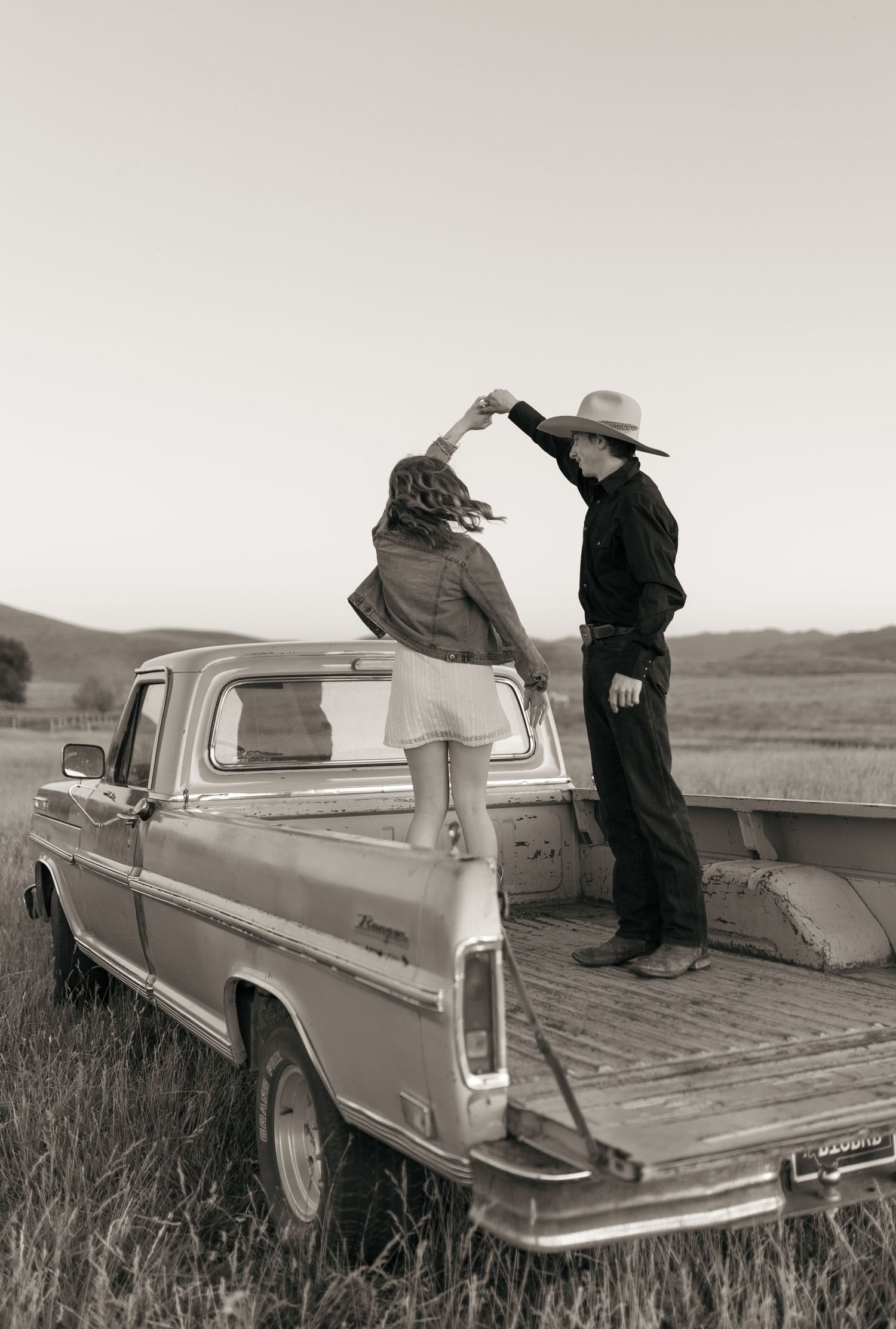A couple sits casually on the bed of a classic truck in a pastoral black and white scene.