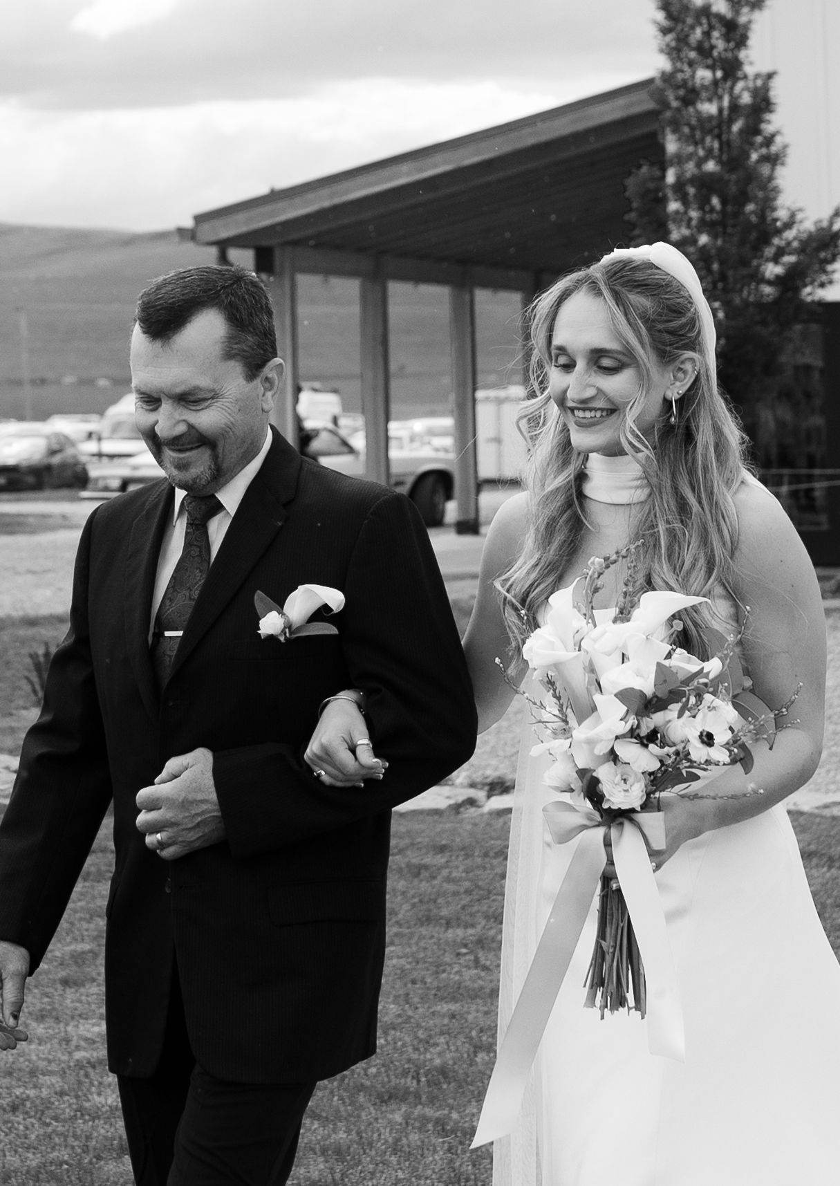 Black and white photo of a smiling bride in white gown holding bouquet, walking arm-in-arm with her dad in wedding in whitefish