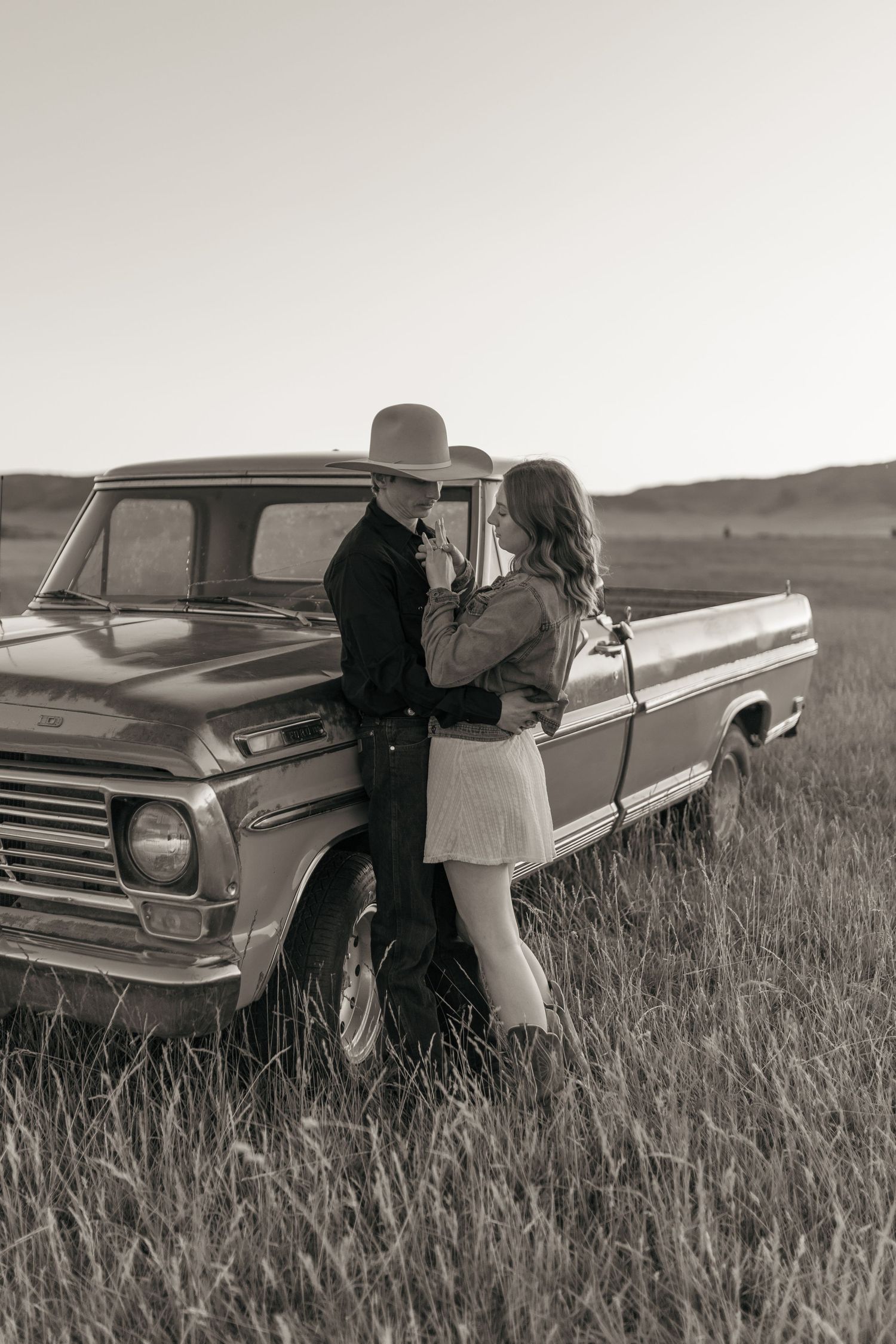 Two people stand beside a classic pickup truck in a serene black and white countryside scene.