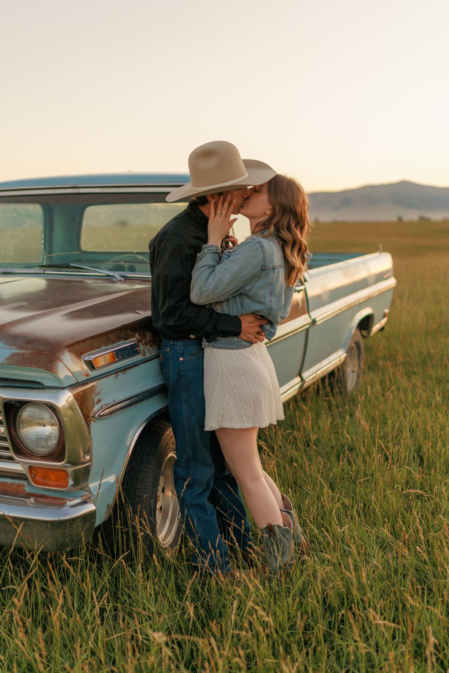 Intimate moment shared between a couple by their classic car in a rural field at dusk.