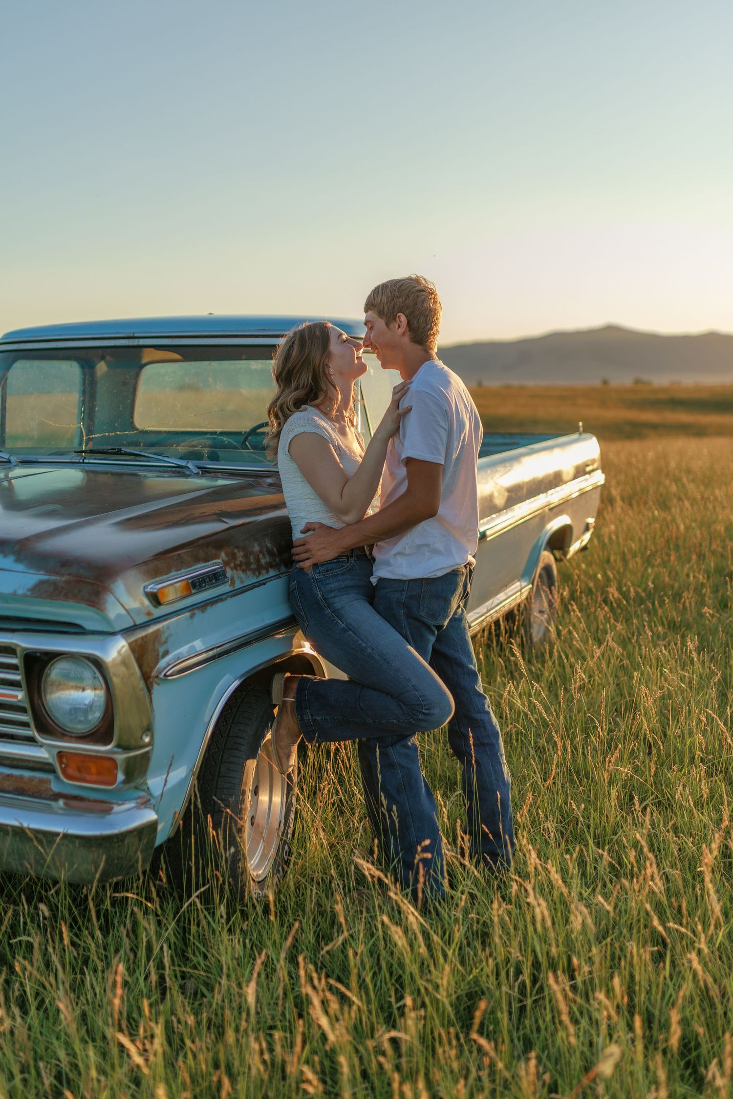 Couple shares a romantic moment leaning against a vintage pickup truck in a golden wheat field at sunset.