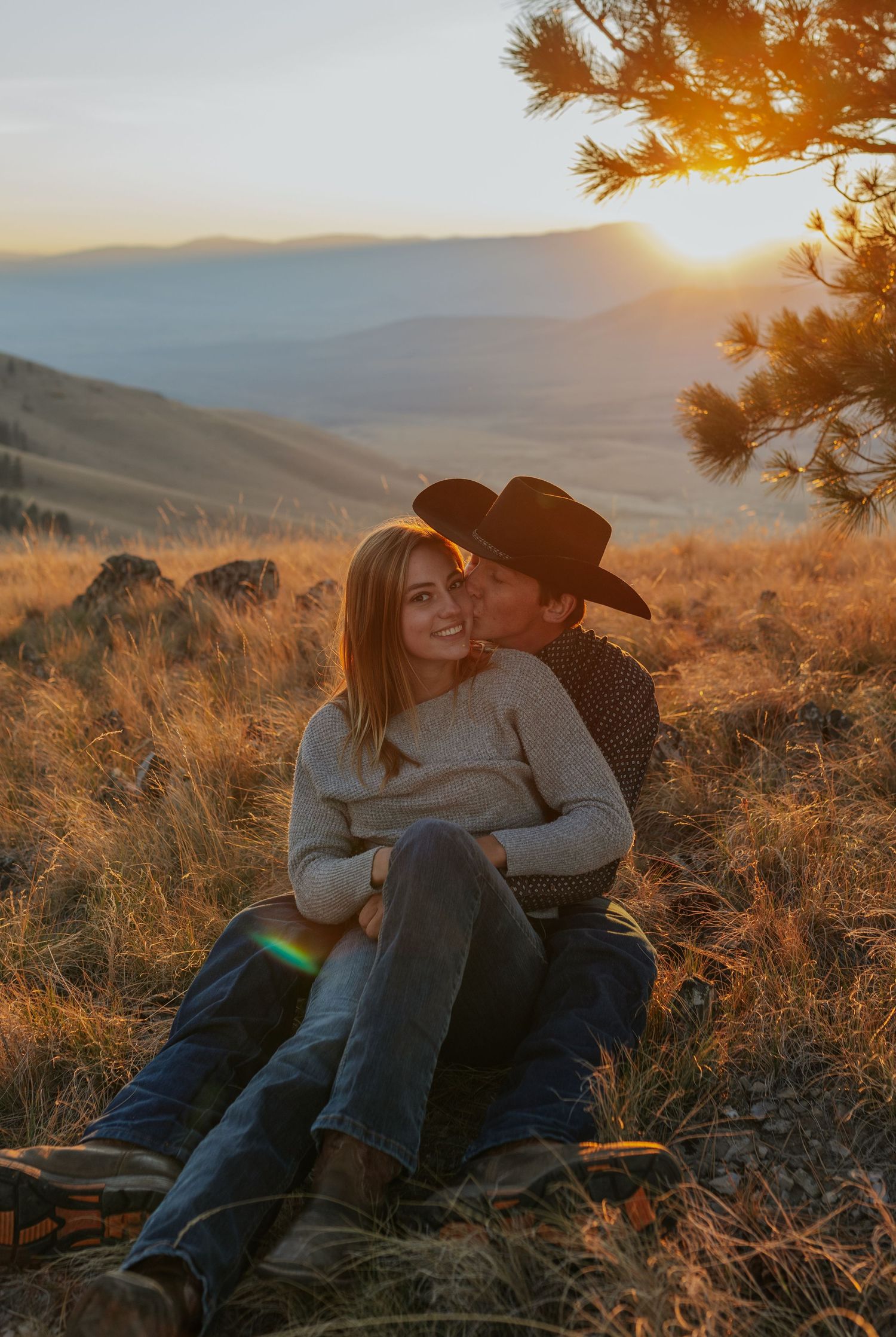 Couple cuddling on grass during sunset with mountains and pine trees creating a romantic backdrop.