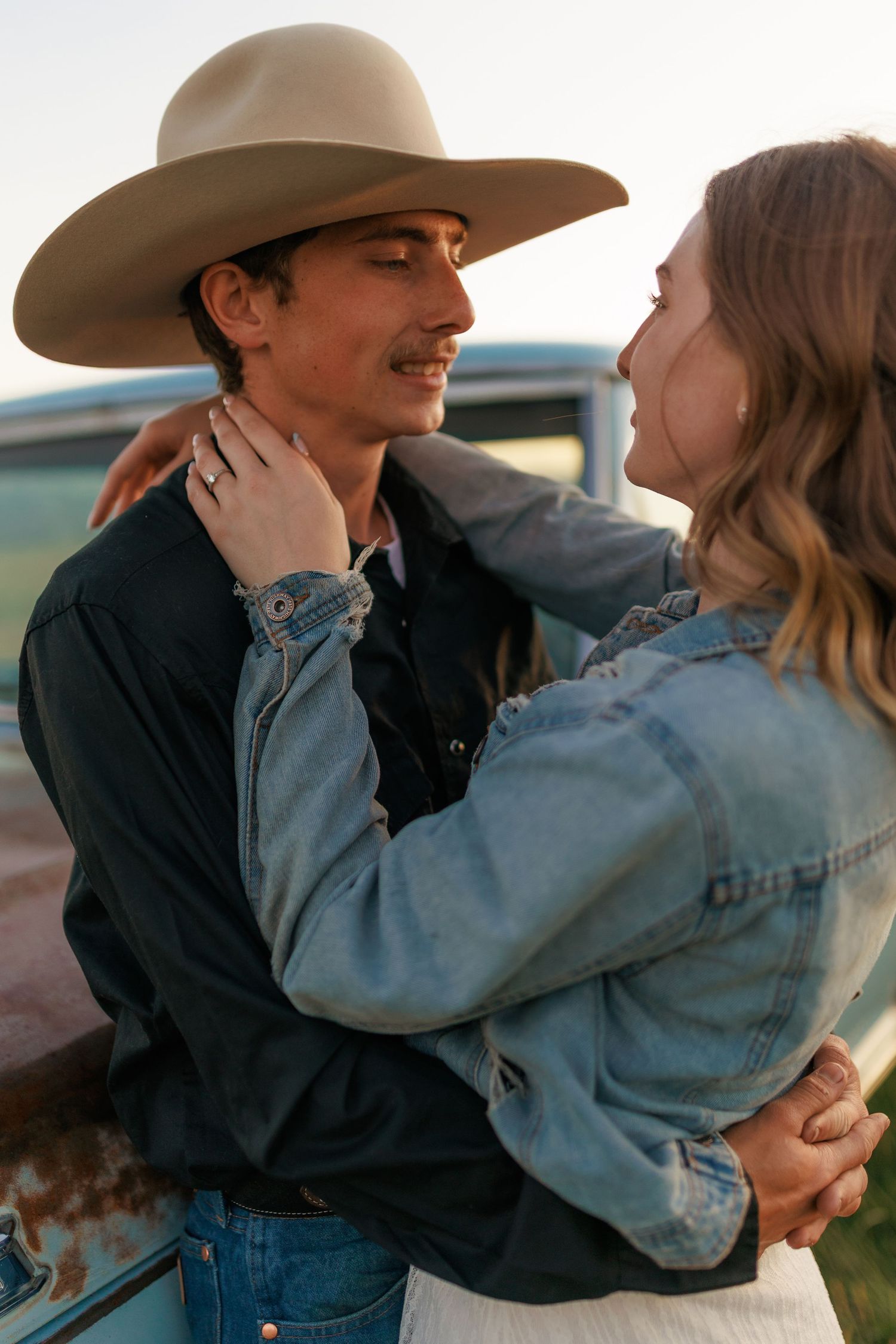 Couple in western attire sharing tender moments against a vintage car backdrop.