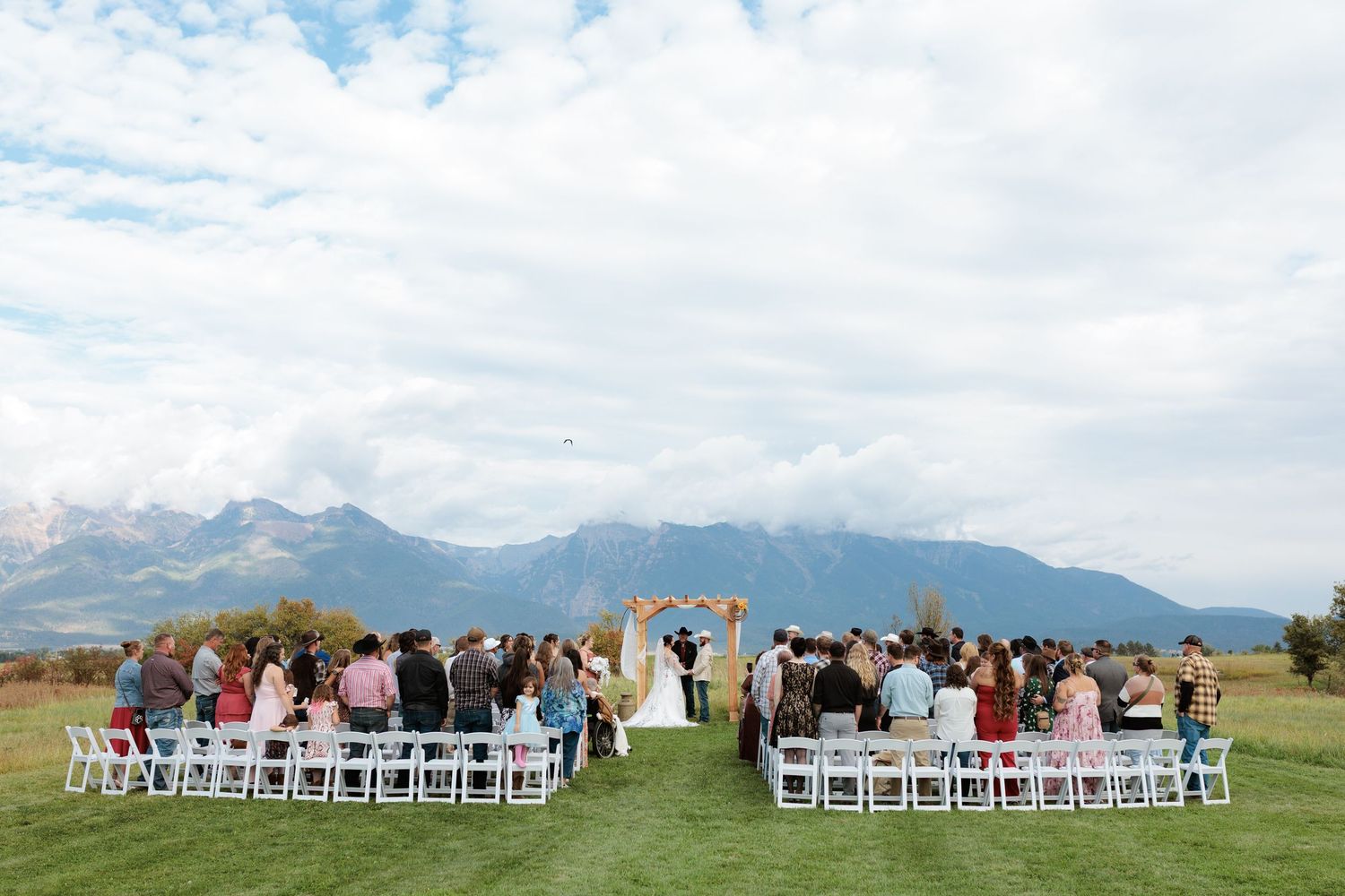 Outdoor wedding ceremony on green lawn with mountain backdrop, white chairs, and floral arch under cloudy sky in Montana.