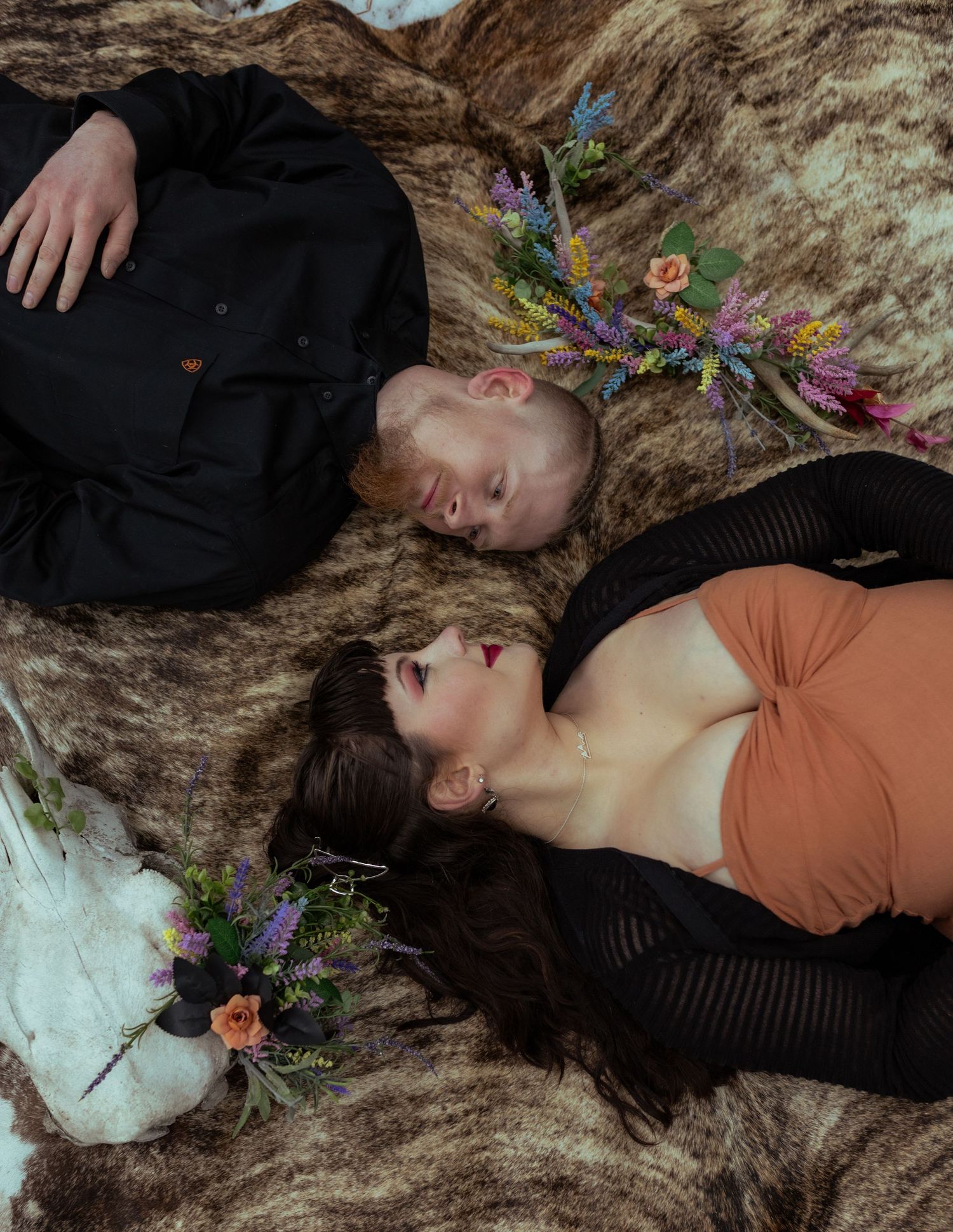 A couple lies on a wooden surface with colorful floral arrangements surrounding them.