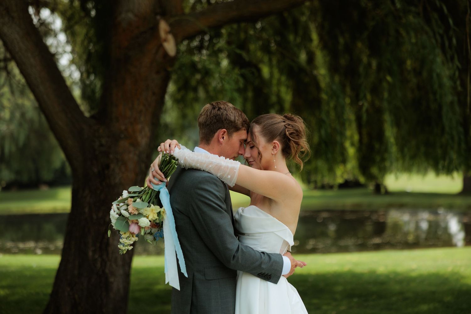 A bride and groom share a romantic forehead touch while embracing outdoors near a willow tree, bride holding a floral bouquet.