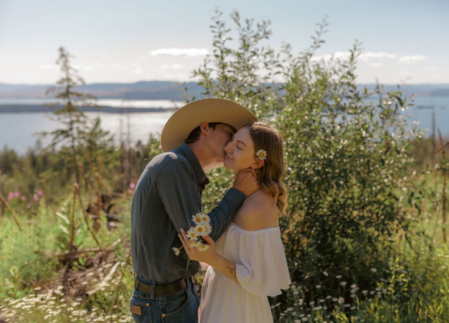 A tender romantic moment unfolds in a sunlit meadow with mountains in the distance.